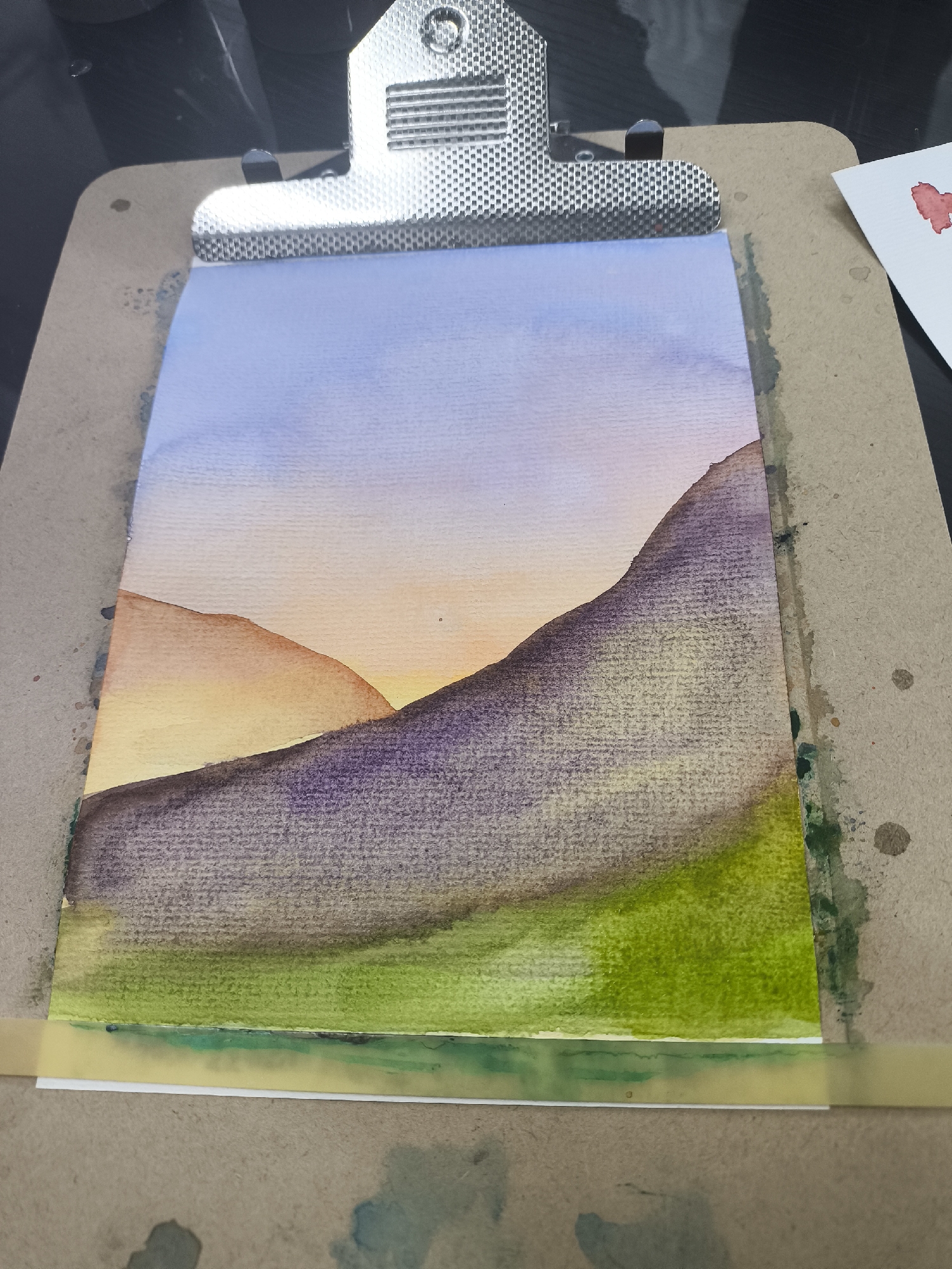 A watercolor painting of a landscape is clipped to a clipboard. The sky is blue and blends into an orange-yellow horizon. Two hills, one orange and the other purple, overlap. The bottom of the painting is green. The clipboard is brown and has paint splotches on it. There is a small white paper with a red paint spot to the right of the clipboard. The background is black.
