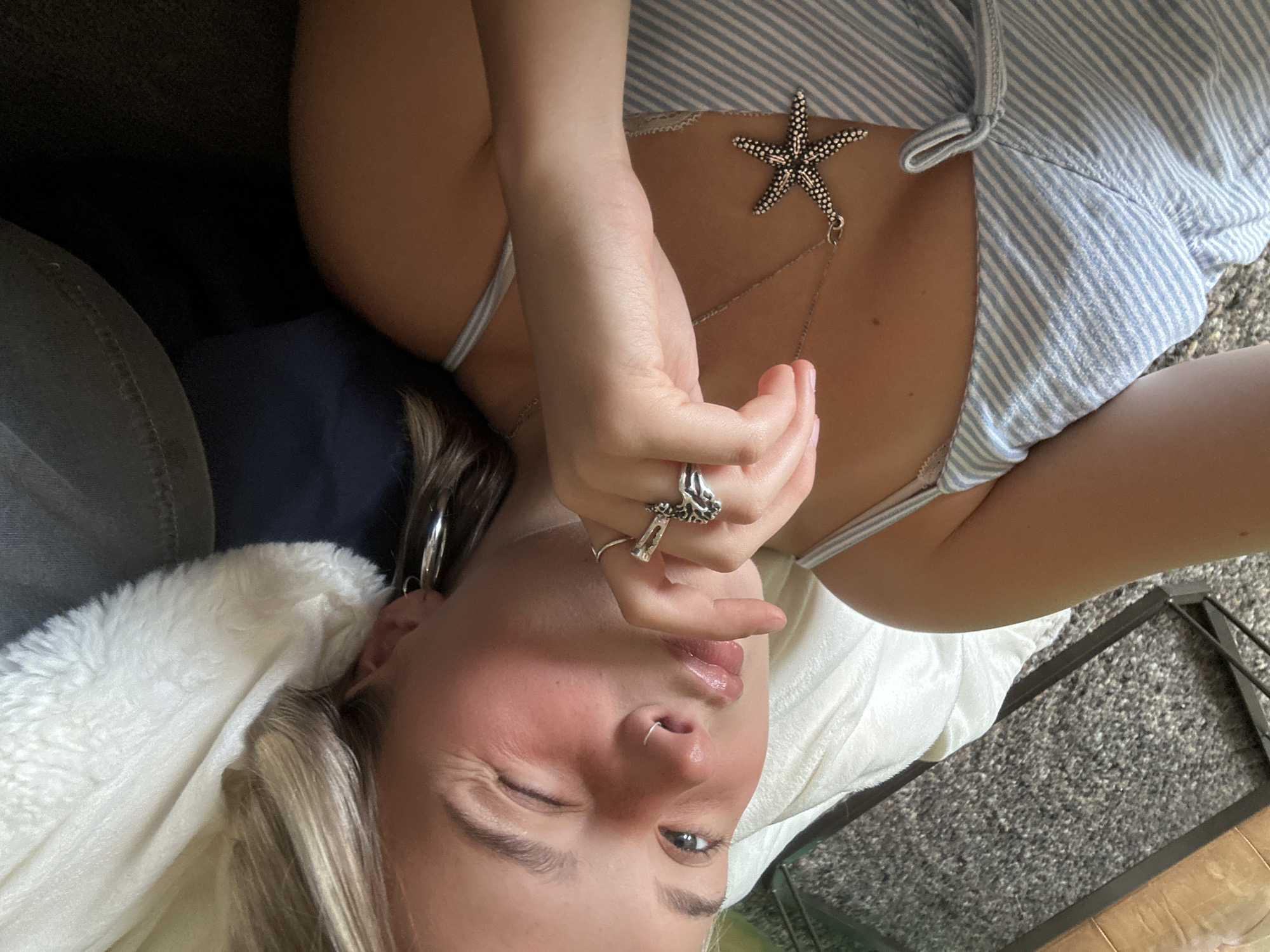 A young, fair-skinned female is depicted in a close-up, high-angle shot, positioned in a reclining posture. Her blonde hair, with lighter highlights, frames her face. Her left eye is open and directed towards the camera, while her right eye is closed in a wink. Her lips are puckered in an expressive pose, with her left index finger lightly touching her lower lip. A small silver ring is visible in her left nostril. She wears a plain silver band on her left index finger and an ornate silver ring on her left middle finger. Silver hoop earrings are present in both ears. A silver chain necklace with a textured, five-pointed starfish pendant rests on her chest. She is wearing a light blue and white striped top with thin shoulder straps and a tie detail at the front. The subject is resting against a white, plush, faux-fur-like material, likely a pillow or blanket, on her left side. A dark blue fabric is visible behind her head. To her right, a dark grey, textured carpet or rug is visible, along with a portion of a dark-framed object. The scene is located in the city of Squamish, Canada.