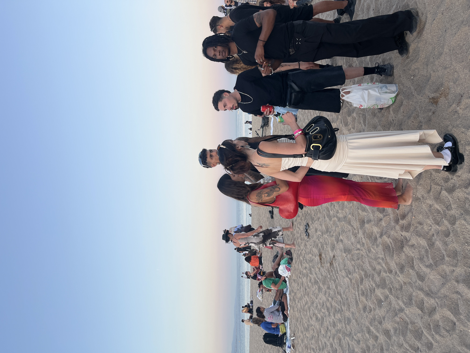 The image depicts a crowded beach scene in Sant Adrià de Besòs, Spain, during late afternoon or early evening. The sky transitions from light blue overhead to a warm, faint pink-orange hue near the horizon where it meets the calm Mediterranean Sea. Distant landforms are visible on the left horizon.
In the immediate foreground and midground, numerous individuals are distributed across the sandy beach. A prominent group of five people stands on the right. This group includes:
1. A woman wearing a light beige, asymmetrical hem skirt and a white top, with dark hair tied back, visible back tattoos, and black Mary Jane style shoes with white socks. She carries a black purse with gold oval embellishments.
2. To her right, a man with short, dark curly hair, wearing a black t-shirt, black shorts, and a black fanny pack. He holds a red beverage can.
3. Further right, a man with dark dreadlocks, dressed in a black t-shirt and black pants with a metallic-accented belt. He wears multiple necklaces with pendants and holds a small item in his right hand.
4. Partially visible on the far left of this standing group, a woman in a sheer, gradient red-to-orange full-length dress holds a red crescent-shaped shoulder bag. She has tattoos on her left arm.
5. Other individuals are visible in the background of this group, including a man in a dark shirt and a cap. A white plastic shopping bag rests on the sand near their feet.
To the left and further back in the midground, several smaller groups and individuals are relaxing on the sand. Some are seated on blankets or directly on the sand, while others stand. Notable figures include:
- A person in a blue shirt seated near a black backpack.
- An individual in a striped shirt conversing with a person in a green t-shirt.
- A person in a white dress and black hat is seen walking from left to right in the midground.
- In the more distant midground, a person is playing a brass instrument, possibly a sousaphone or tuba.
The sand is light brown and shows numerous footprints. Various personal items like sandals, backpacks, and beverage cans are scattered on the beach. The overall impression is one of a social gathering or leisure activity on the beach as the day transitions to evening.