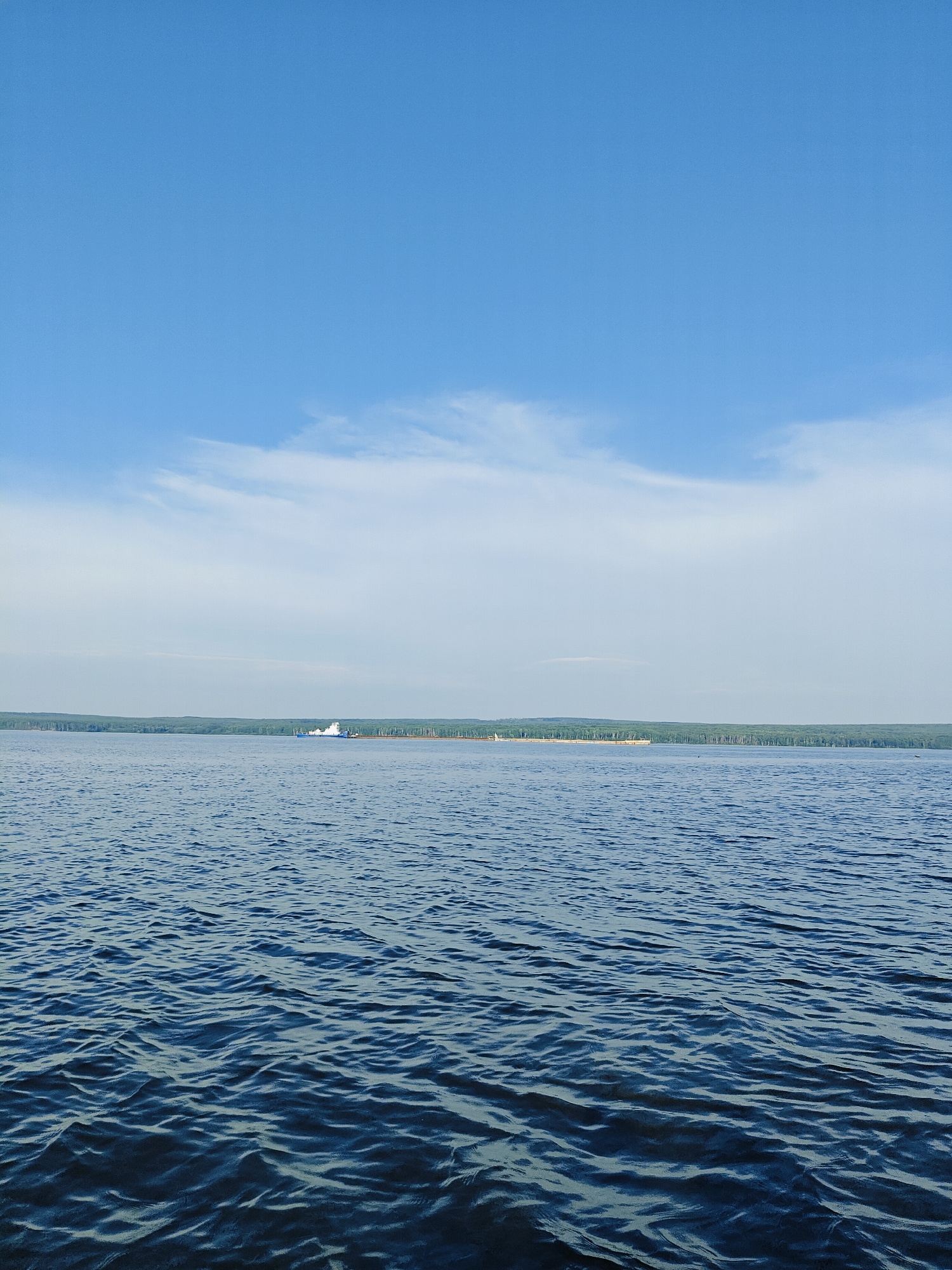 The image depicts a wide expanse of blue water, likely a river or lake, under a clear blue sky with wispy white clouds. The water is calm with gentle ripples. In the distance, a dense line of green trees forms the shoreline of what appears to be a landmass. On the water, near the distant shore, there are several white structures, possibly boats or buoys, and a few scattered trees are visible along the coastline. The scene is serene and natural.
Based on the visual elements of a wide body of water bordered by a dense forest and the presence of what could be a riverine or lakeside settlement, this scene is consistent with the environment around the city of Chaikovsky, Russia, which is situated on the Kama River. The city is known for its industrial significance and its location on this large river.
