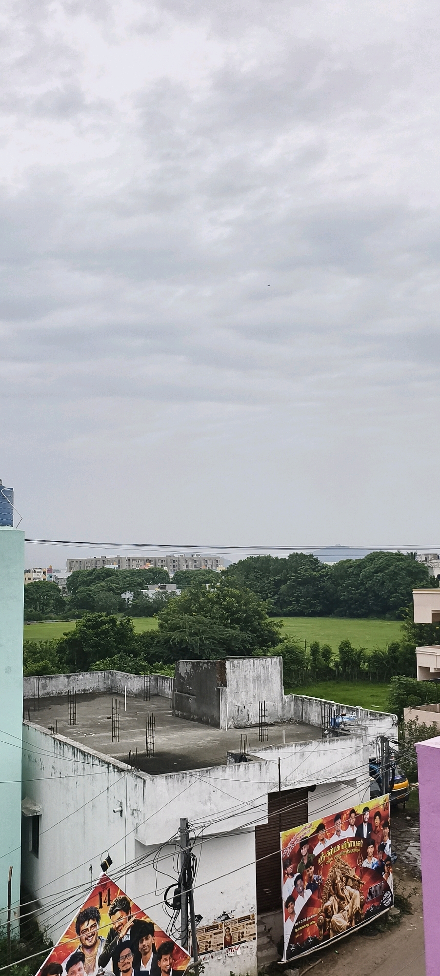 An overcast sky looms above a dense canopy of green trees, suggesting a humid day in Kundrathur, India. The scene is dominated by a broad expanse of green lawn, possibly a park or open field, bordered by various structures. In the foreground, a partially constructed concrete rooftop is visible, with exposed rebar suggesting ongoing construction or disrepair. A light blue building is partially visible on the left edge of the frame. A prominent billboard advertising a film featuring multiple actors is affixed to a wall in the lower right quadrant. Behind the billboard, a portion of a small yellow vehicle, possibly an auto-rickshaw, is visible. In the distance, a large, multi-story building complex with a light-colored facade stands amidst the trees, hinting at a developing urban area. The overall impression is one of a typical Indian cityscape, balancing natural elements with the presence of human habitation and commerce.