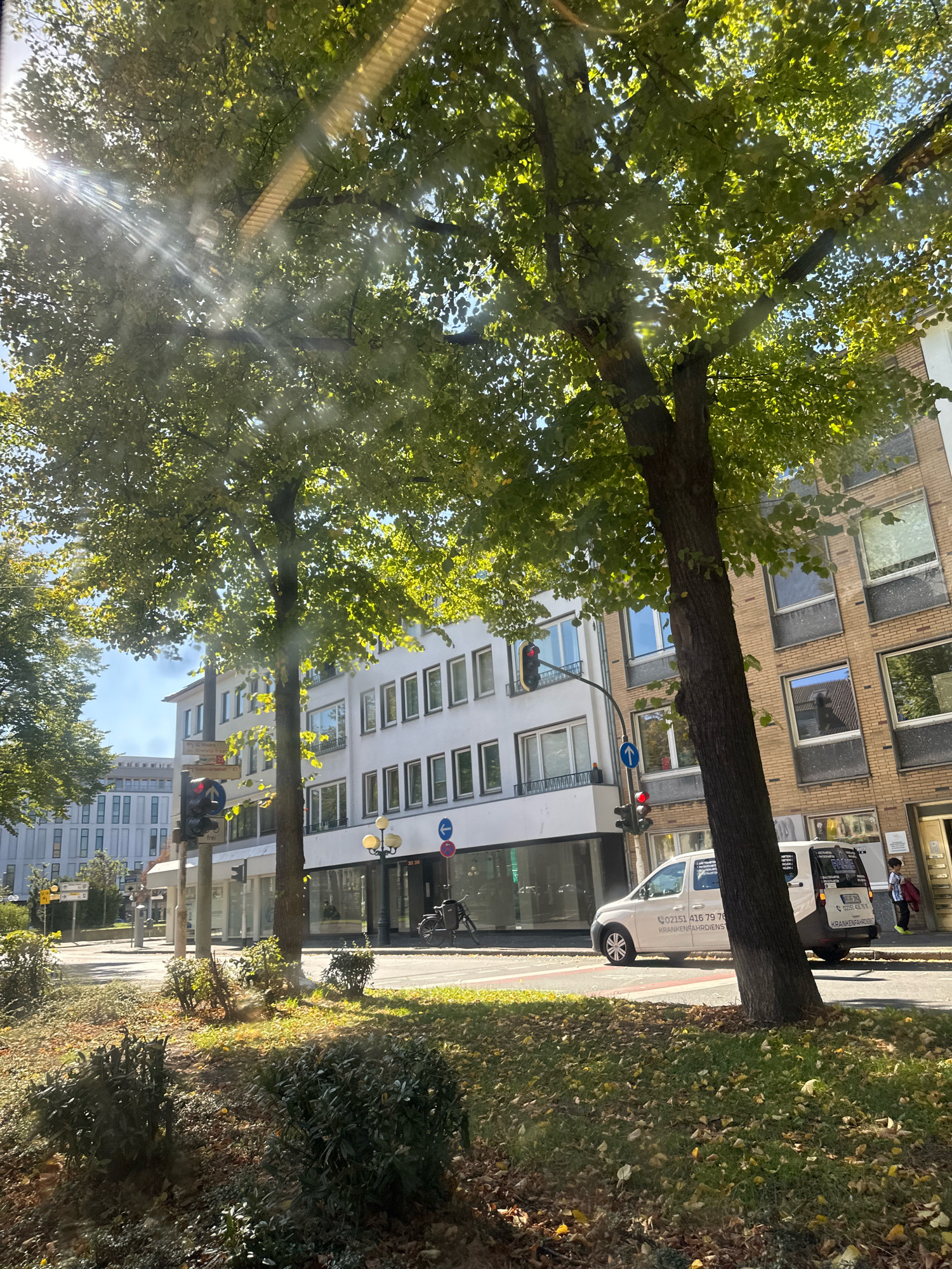 This is a ground-level, daytime, outdoor photograph of a street scene in Krefeld, Germany, as indicated by the text on a signpost and a vehicle. The image captures a tree in the foreground, with its branches and leaves partially obscuring the view of buildings and the street. Sunlight filters through the leaves, creating lens flare and dappled light effects.
On the street, a white van is parked, with visible text on its side, "02151 416 79 76 KRANKENFAHRDIENST". A traffic light with a red signal is visible to the left of the van. Further down the street, several buildings are present, characterized by their white and brick facades and numerous windows. A person wearing a blue and white jacket and bright green shoes is standing near the entrance of one of the buildings. In the background, more buildings and trees are visible. The ground is covered with grass and fallen leaves, suggesting it might be autumn.
The signpost displays German text and symbols, including directional arrows and a street name, "Schulstr. Krefeld-Traar", which firmly places the location in Krefeld, Germany. The overall atmosphere is that of a quiet urban street on a sunny day.
