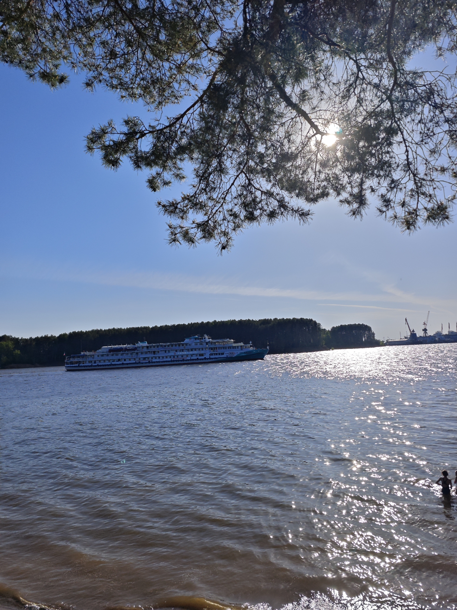 The image captures a serene scene on the Kama River in Chaikovsky, Russia. A large, multi-decked passenger ship glides across the river, positioned to the left of the frame. A dense forest lines the opposite bank. To the right, industrial cranes are visible near a pier. Sunlight glints off the water's surface, creating a shimmering effect. In the lower right corner of the frame, two individuals stand in the shallow water. Branches of a coniferous tree overhang the top of the image, partially obscuring the bright sun. The sky is blue with a few scattered clouds.