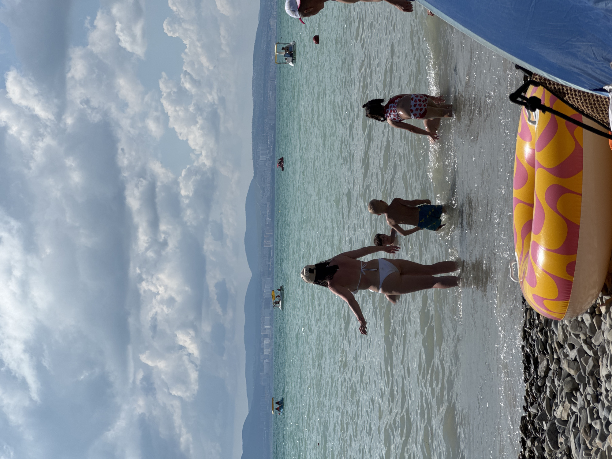 In this vertical, eye-level shot, several people are enjoying a sunny day at the beach in Novorossiysk, Russia. The scene features a body of water with gentle waves, transitioning to a pebble-strewn shoreline. In the foreground, a large, inflatable ring with a yellow and pink swirl pattern rests on the beach.
Several individuals are in the water. A woman in a white bikini stands with her arms outstretched, facing the water. Further out, a child in red and white polka dot swimwear is crouched down. A young boy in blue shorts is also in the water, facing away from the camera. In the background, several small boats or watercraft with people are visible on the lake. The distant shoreline is marked by a city skyline backed by rolling hills or mountains.
The sky above is partly cloudy, with a mix of bright white clouds and areas of blue sky, suggesting it's daytime, likely late morning or early afternoon. The water appears clear and a light turquoise color. The overall mood is one of leisure and recreation, with people enjoying a summer day by the water. No visible text is present in the image.
