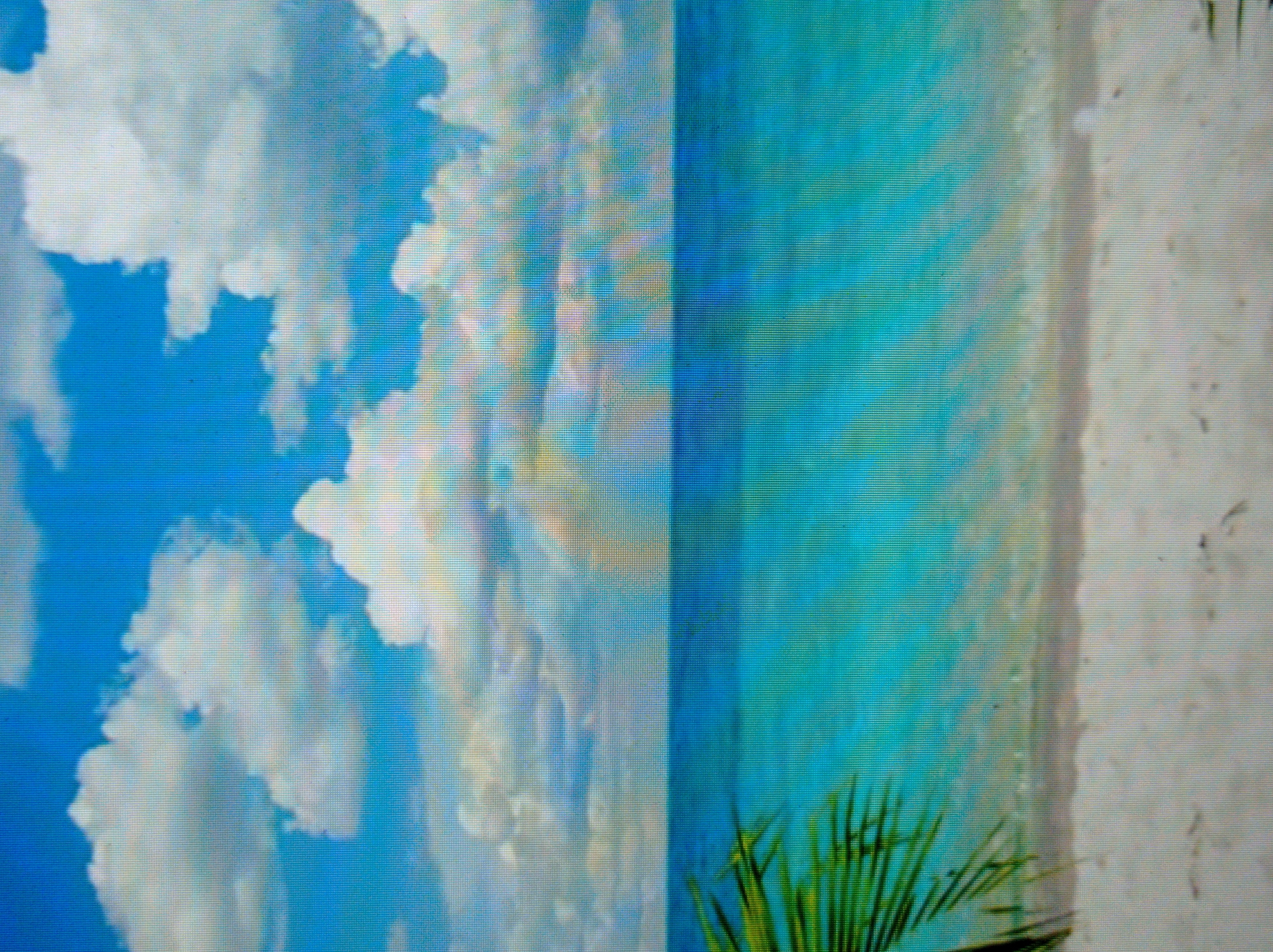 The image depicts a tropical beach scene. The upper portion of the image features a bright blue sky with scattered white clouds. Below the sky is the ocean, rendered in vibrant shades of turquoise and blue, suggesting clear, tropical waters. The horizon line separates the sky and sea. In the foreground, there is a sandy beach, appearing light in color. On the left side of the image, a portion of a green palm frond is visible, extending into the frame from the edge. The image does not contain any people, specific landmarks of Mukachevo, Ukraine, or any discernible actions or interactions. It is a generic representation of a beach.
