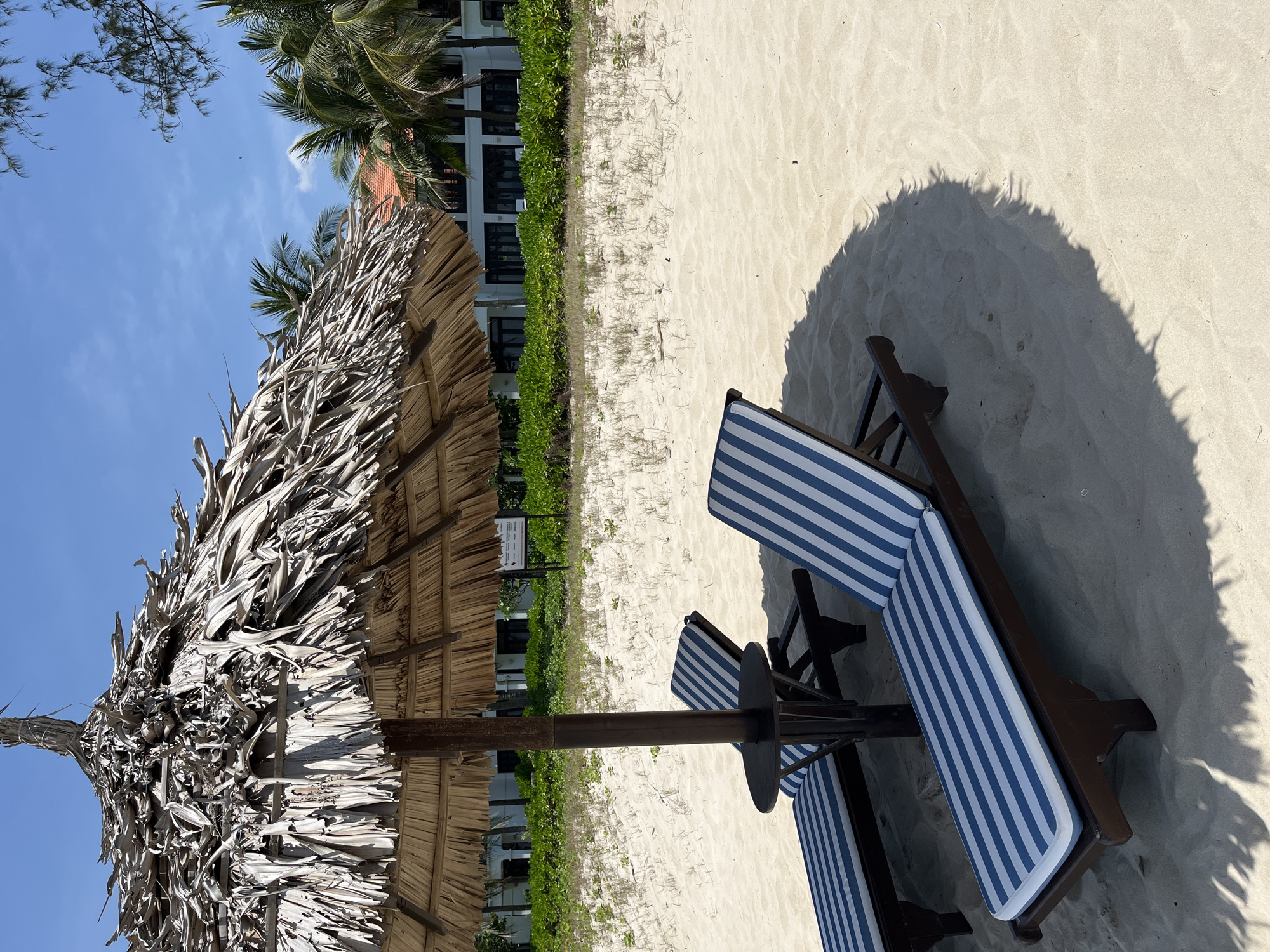 The image shows a beach scene in Đà Nẵng, Vietnam. A thatched-roof beach umbrella provides shade for two blue and white striped beach chairs on the sandy beach. The shadow of the umbrella and chairs is cast on the sand. In the background, there's a building with green vegetation in front of it. Palm trees are visible above the building and to the left of the umbrella. The sky is blue with some clouds.