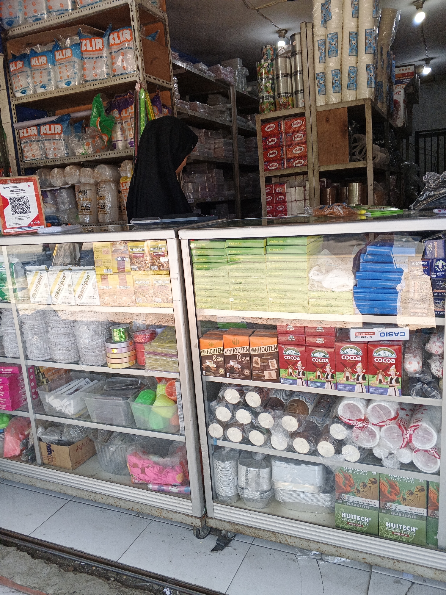 This is a medium shot, eye-level interior photograph of a small retail shop selling various disposable goods. The shop is filled with shelves packed with products like paper cups, food packaging, and other household items. In the foreground, a glass-fronted display cabinet showcases rows of these items. A woman wearing a black hijab is visible behind the counter, partially obscuring some of the products on the shelves. The lighting appears to be a mix of fluorescent lights from the ceiling and possibly natural light from the entrance, creating a well-lit but somewhat cluttered environment. The overall impression is of a busy, local convenience store or variety shop. This shop is located in Sukabumi, Indonesia.