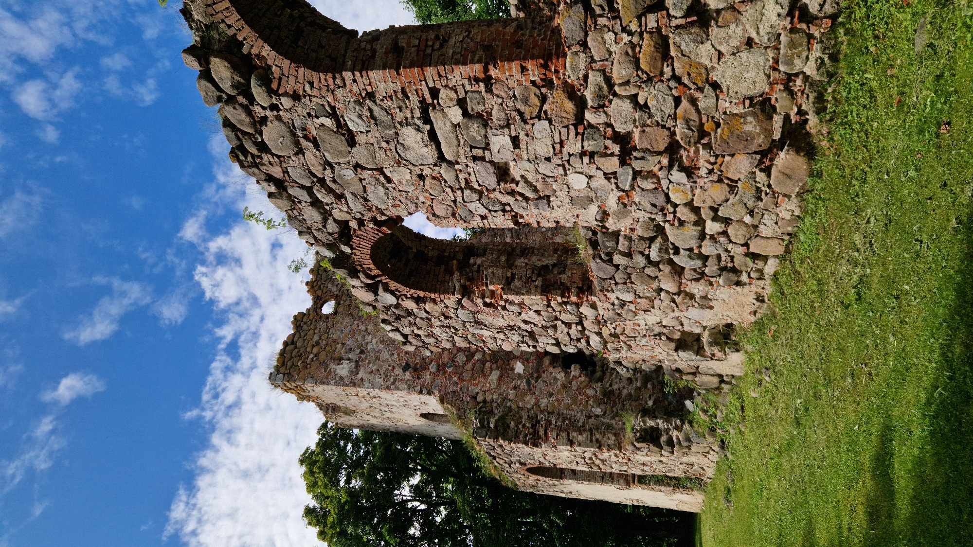 A low-angle, full shot captures the weathered stone ruins of the Embūte Evangelical Lutheran Church against a vibrant blue sky dotted with scattered white clouds. The ancient structure is composed of large, irregularly shaped stones and red brick, with arched openings visible, hinting at its former grandeur. Green grass covers the ground in the foreground, with a large, dark green tree on the left side of the frame. The scene is bathed in bright sunlight, casting shadows on the grass and highlighting the textures of the stonework.