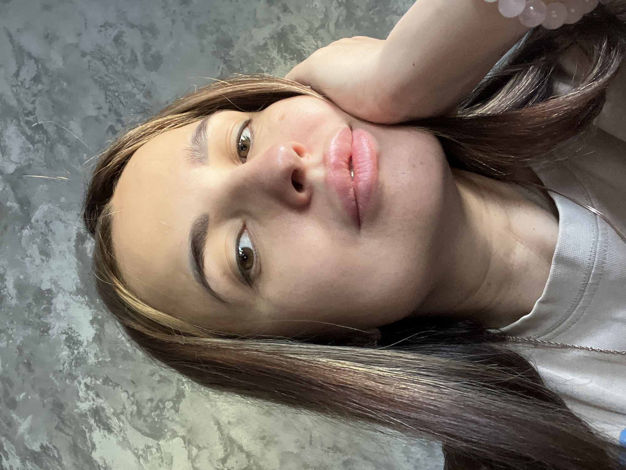 A close-up, selfie-style portrait shows a woman with long, brown hair with highlights. She is looking directly at the camera, holding her face with one hand, which has a bracelet. Her eyes are brown, and her lips appear full. She is wearing a white shirt, and a thin silver necklace is visible. The background is a gray, mottled surface. The location is unknown.