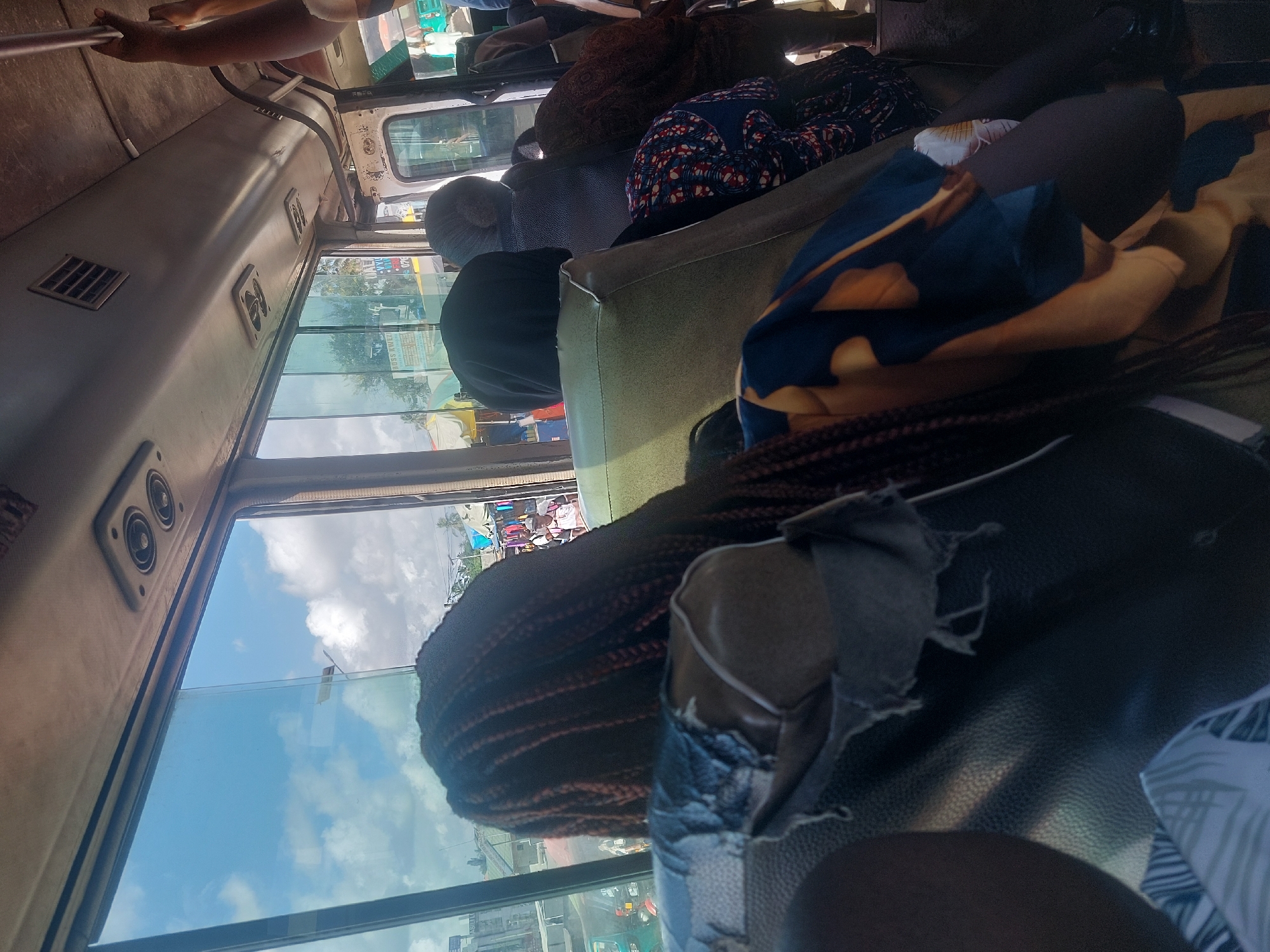 The image depicts the interior of a public transport vehicle, likely a bus or mini-bus, seen from a passenger's perspective looking forward and towards the side windows. The overall scene suggests a busy daytime journey in an urban environment.
In the foreground, the braided hair of a passenger is visible, along with their shoulder and part of a dark top. The seat in front of this perspective is upholstered in worn black material. Further inside the vehicle, several other passengers are visible. One person wears a black top and a grey beanie or head covering. Various patterned fabrics, including blue with white and red accents, and a blue and tan/yellow design, are seen on or draped over seats. The vehicle's ceiling features overhead panels with vents or small fans, and a silver handrail is present.
Outside the vehicle, a vibrant street scene unfolds through the large side windows. The sky is bright with patches of blue and white clouds, indicating daytime and fair weather. Buildings, trees, and what appear to be market stalls or shops with colorful tarps and awnings line the street. Other vehicles are also visible, suggesting active traffic.
The activity is people commuting or traveling by public transport. No strong emotions are discernible among the passengers; they appear to be engaged in routine travel. While the setting aligns with an urban location like Dar es-Salaam, Tanzania, there is no clearly legible text on any visible signs or objects to definitively confirm the location within the image itself. The vehicle's interior shows signs of age and regular use.