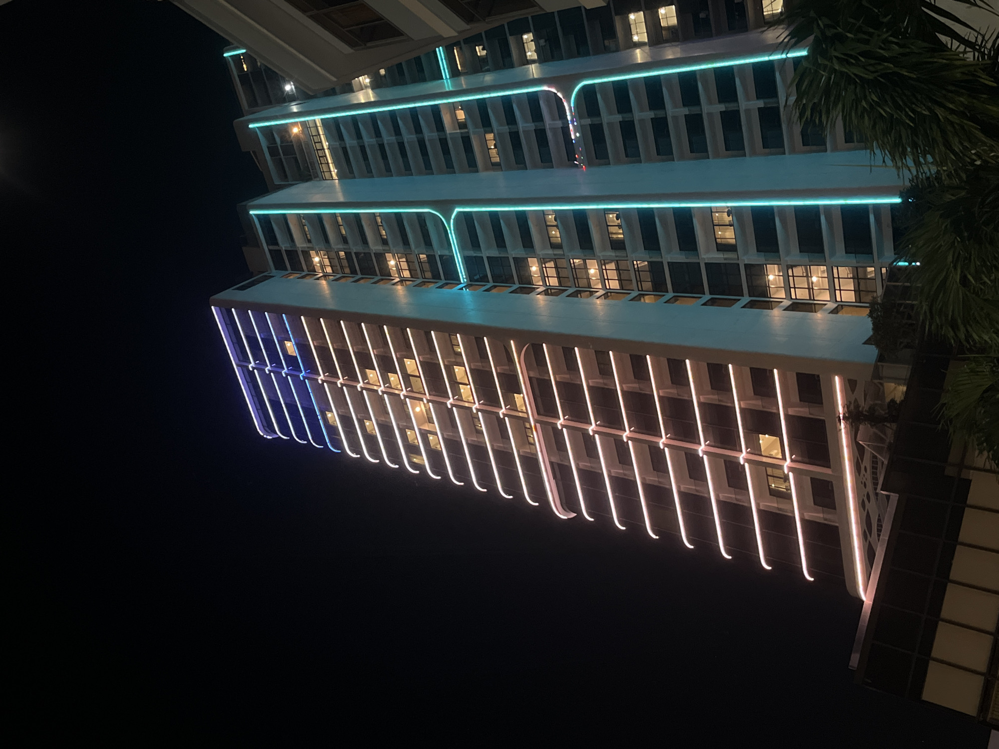 The image captures a nighttime view of a modern building in Bac Nha Trang Ward, Vietnam. The building is illuminated with colored lights, transitioning from blue at the bottom to pink towards the top. Horizontal lines of green neon lights delineate the different levels of the building. The dark sky provides a stark contrast to the bright building. Some palm tree branches are visible on the right side of the image. The building has multiple windows with lights visible inside.