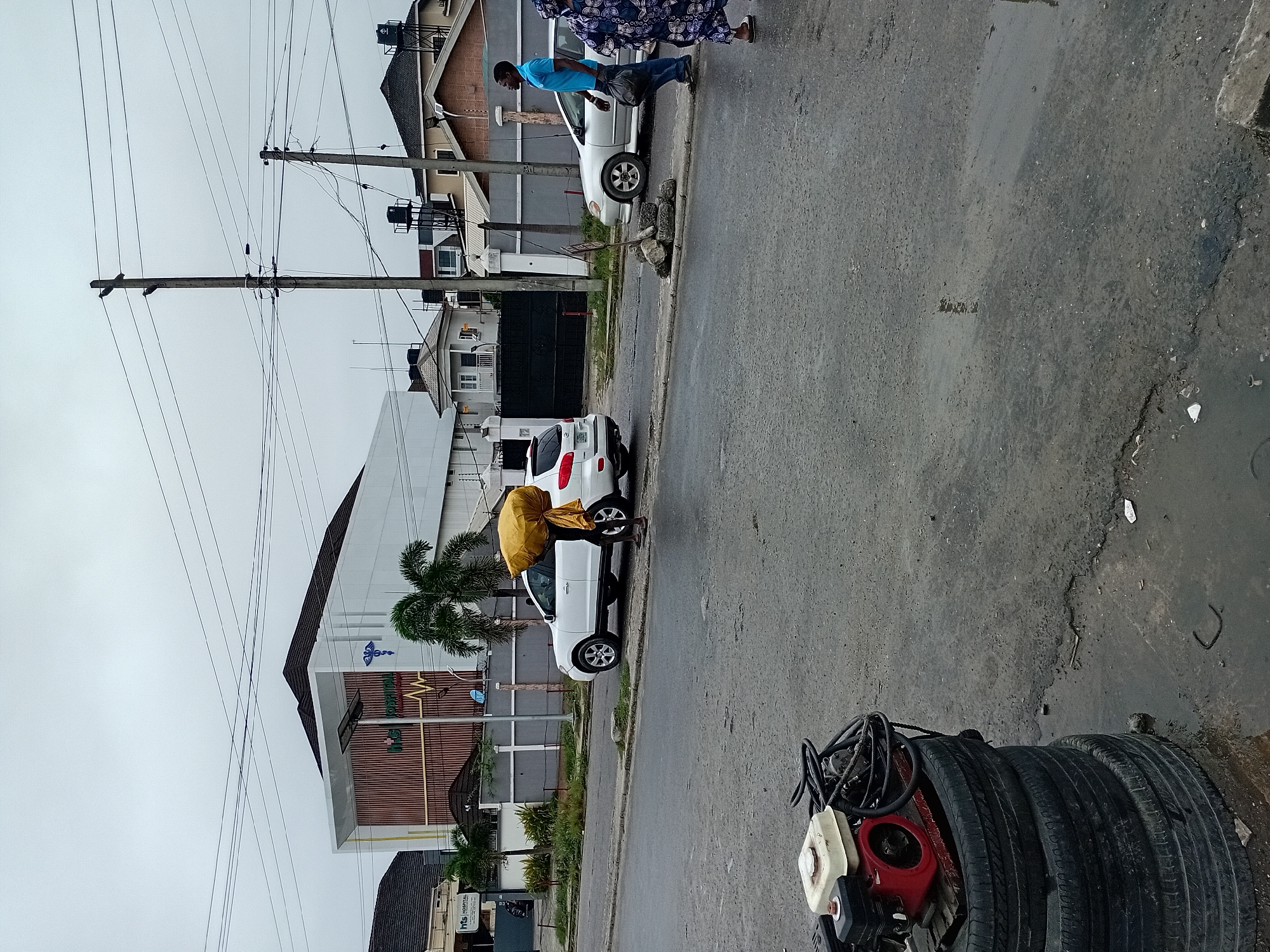 In an outdoor urban scene, a man carries a large, yellow, cloth-wrapped bundle on his head. He is walking on a paved road next to a white SUV. In the background, a modern building with a white facade and vertical wooden accents stands behind a gray fence. Other buildings, including houses with dark roofs and one with a prominent water tank, are visible further in the background. Several utility poles with numerous wires crisscross the sky, which is overcast. In the foreground, a stack of tires sits next to a small engine-powered generator. To the right, a second man in a blue shirt and jeans walks away from the camera, carrying a black bag, and a woman in a patterned blue and white dress walks towards the camera. The road surface appears wet and reflective in places, suggesting recent rain.