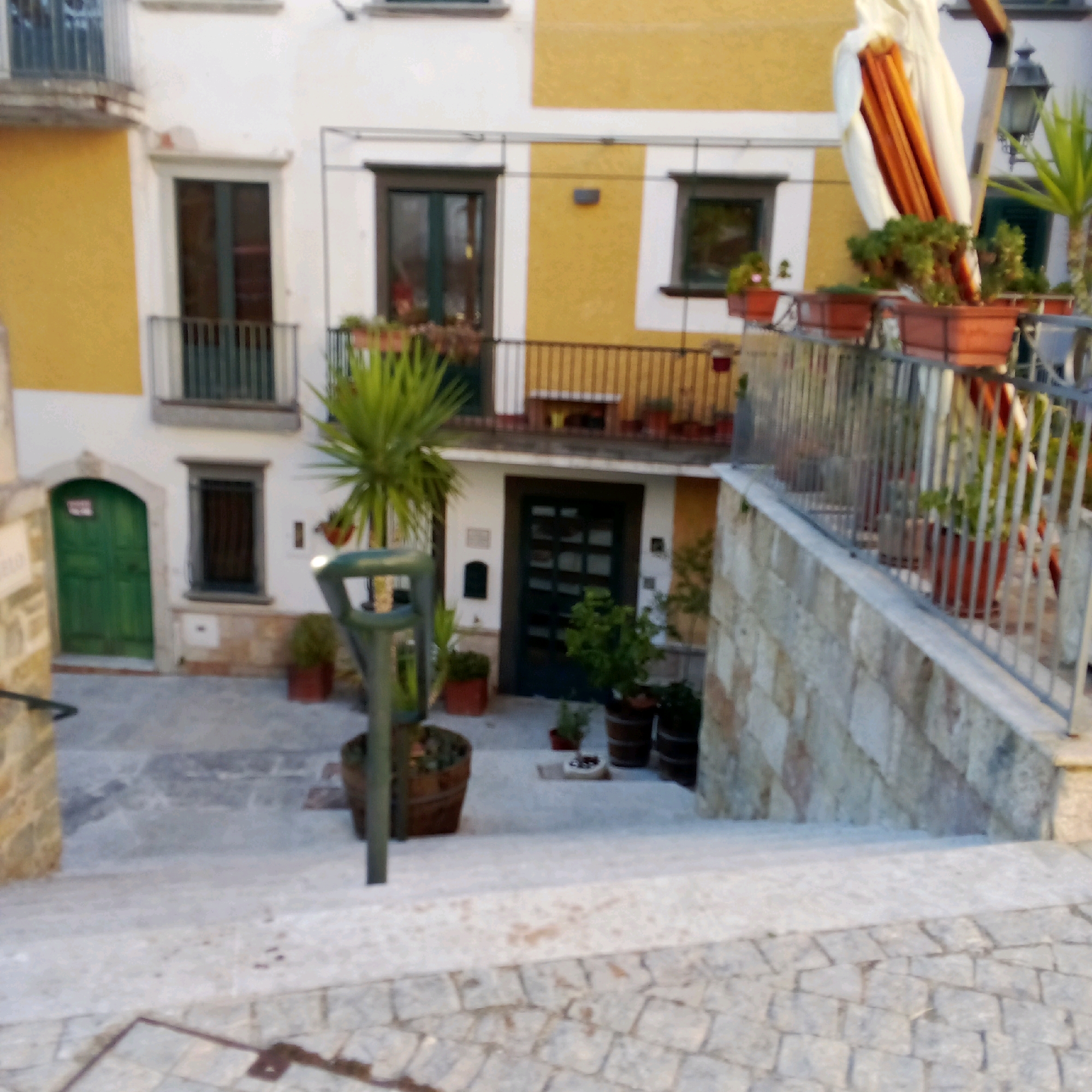 The image depicts a narrow street or courtyard area in the town of Castelnuovo di Conza, Italy. The buildings are made of stone and plaster, with yellow and white facades. There are several doors and windows, some with balconies. Potted plants and trees are visible throughout the scene, adding a touch of greenery. A metal railing lines a staircase that leads up to a building. In the foreground, a cobblestone pathway is visible. The overall impression is one of a quaint and well-maintained European village street.