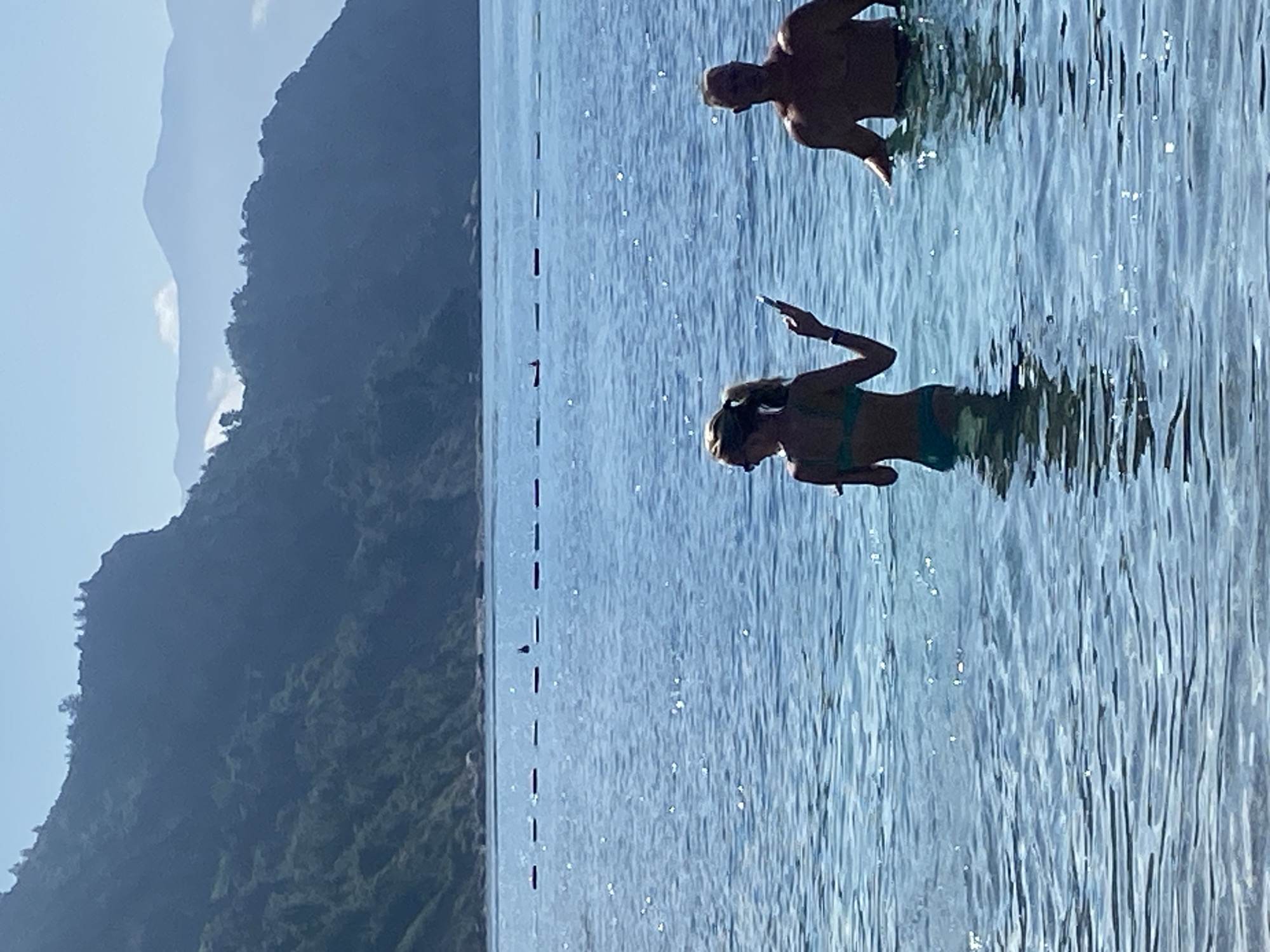 In Prijevor, Montenegro, two people stand in clear blue water, with a mountainous backdrop. A woman in a teal bikini faces away from the camera, holding a smartphone in her left hand. To her right, a man, shirtless, stands with his arms resting on the surface of the water. The water is calm with sun glints and a buoy line stretches across the middle ground. The distant mountains are layered in shades of blue, with a clear sky above.