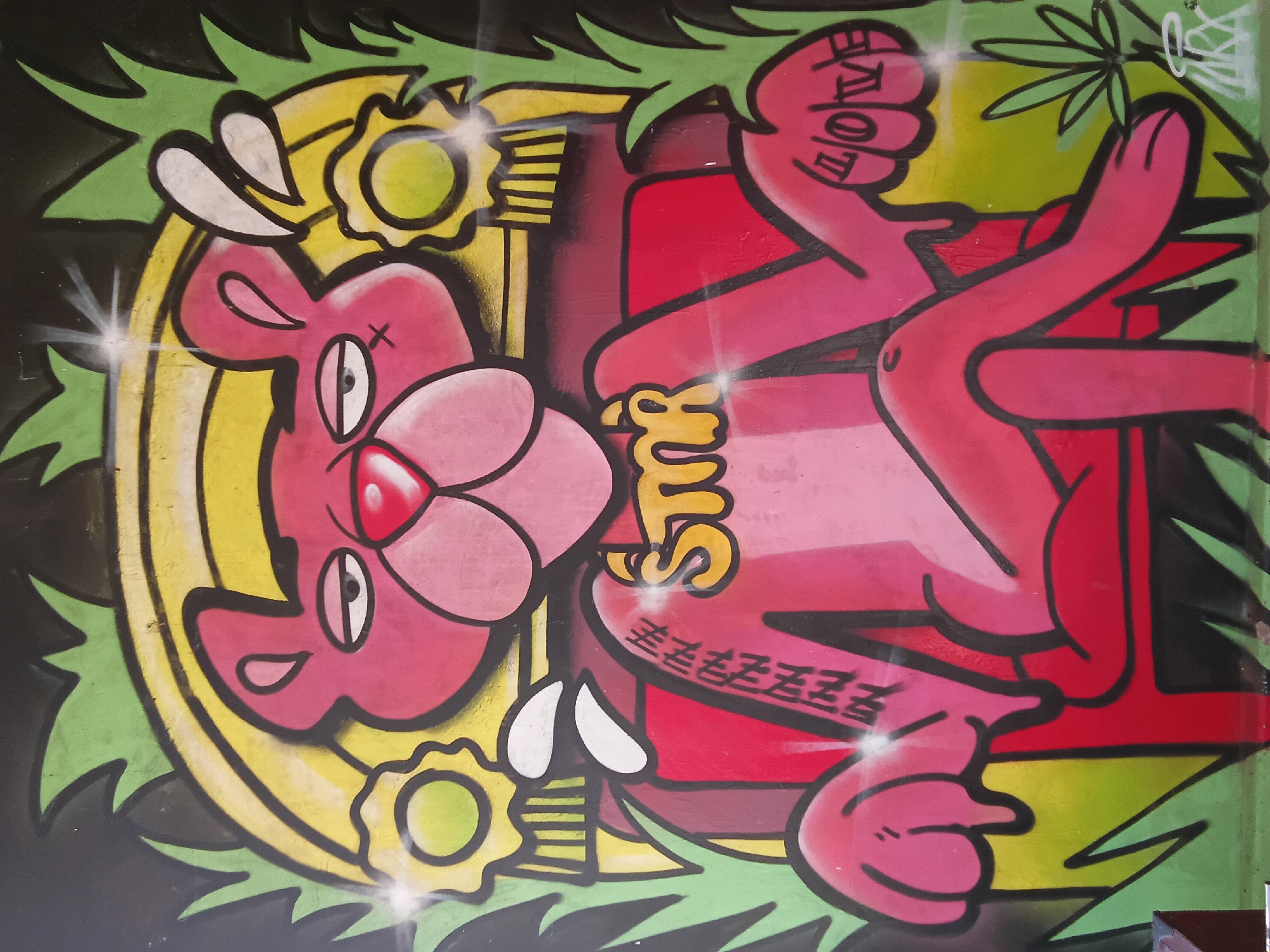 The image shows graffiti art on a wall in Vagator, India. It features a cartoonish, pink panther-like character wearing a gold chain with the text "Stik" and another "Love" on its hand. The character is surrounded by a yellow circle and green, leaf-like shapes. The wall also has some writing in black ink. The background is dark, appearing to be night or dim lighting.