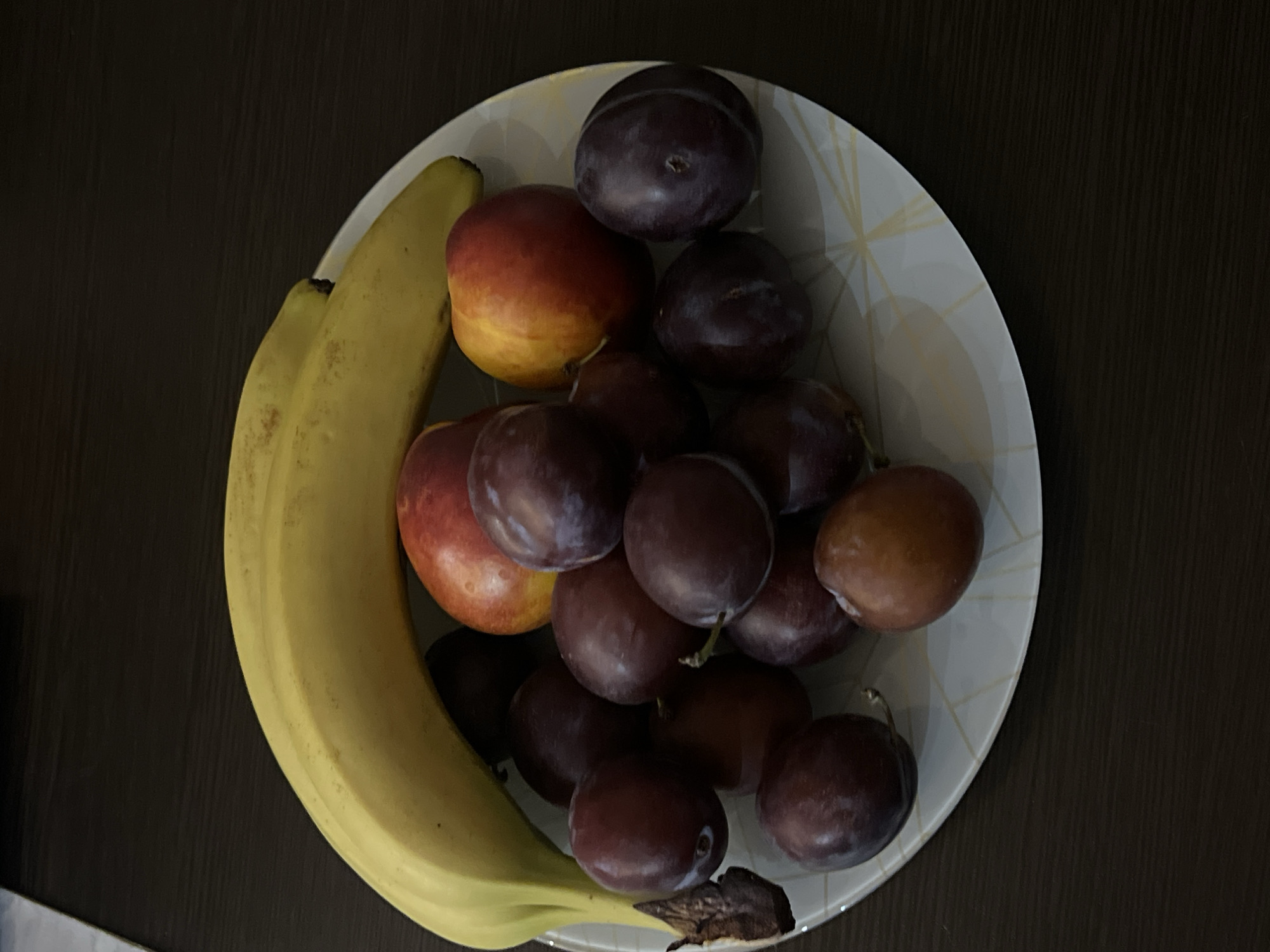 A still life photograph shows a white plate with a geometric pattern holding a selection of fruits: two bananas, several dark purple plums, and two peaches or nectarines. The plate is positioned on a dark brown, horizontally-grained surface. The lighting suggests an indoor setting, potentially a table. The image does not contain any information related to the city of Нежинка, Russia.