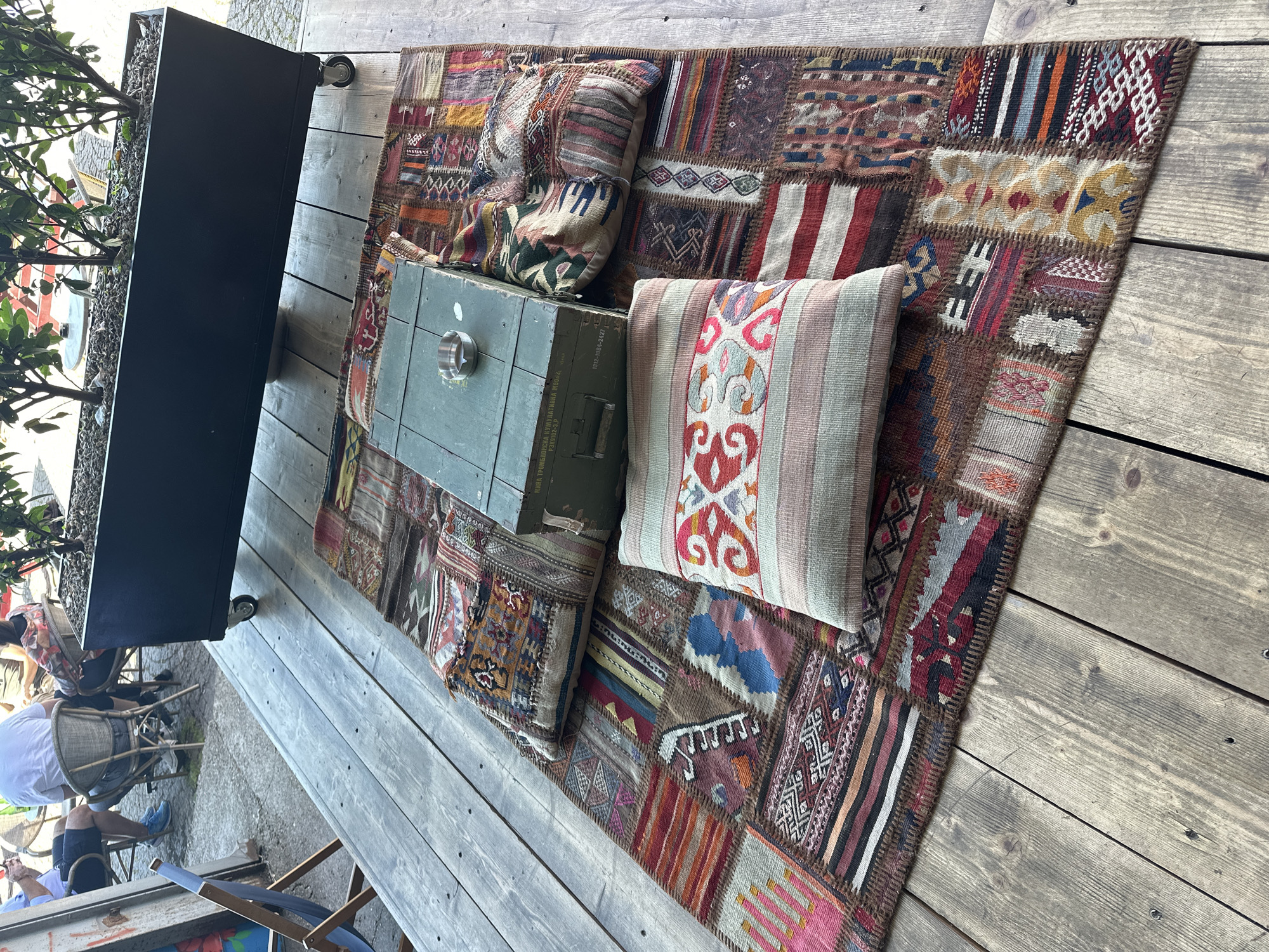 In an outdoor seating area with weathered wooden floorboards in Belgrade, Serbia, a rustic scene unfolds. A large, dark planter box with live plants sits elevated on casters. In front of it, a colorful, patchwork rug is laid out. Two lumbar pillows are placed on the rug. The pillow on the left features a geometric pattern in red, orange, and white against a pale background, bordered by thin stripes of various colors. The pillow on the right displays a more abstract pattern with red, cream, and green hues. A vintage olive-green metal ammunition box rests on the rug, its lid open, revealing a small metal ashtray. In the background, blurred figures are seated at outdoor tables, enjoying the ambiance of the location.