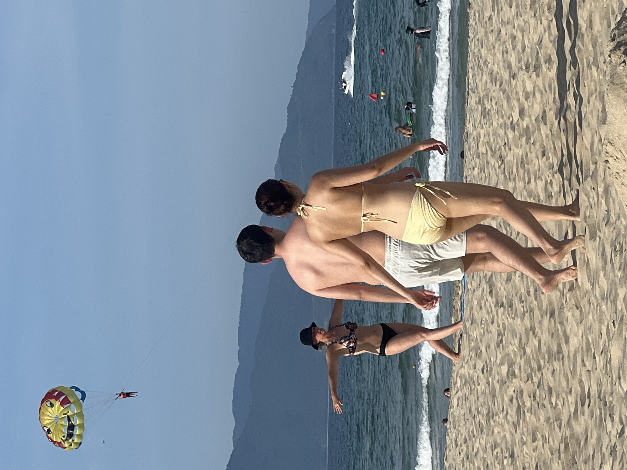 In Đà Nẵng, Vietnam, this image captures a sunny day at the beach, likely My Khe Beach, renowned for its sandy shores and clear waters. In the foreground, a couple walks hand-in-hand away from the camera, the man wearing white shorts and the woman a yellow bikini. Further down the beach, another woman in a black bikini and hat stands with her arms outstretched, facing the sea. In the distance, a paraglider with a smiley face design soars in the bright blue sky, and a jet ski zips across the turquoise ocean. The background features rolling green hills and mountains, adding to the scenic coastal landscape. The sea is calm with gentle waves lapping at the shore, and a few other beachgoers are visible in the water.