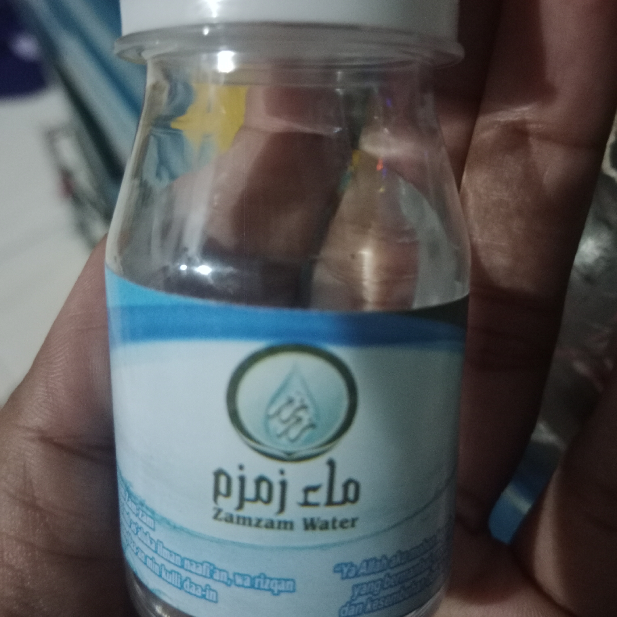 A person's hand is holding a small, clear plastic bottle filled with Zamzam water. The bottle has a white cap and a label with blue and white elements. The label features Arabic script and the text "Zamzam Water" in English. Some additional text in what appears to be Indonesian is also visible on the label. The background is blurred and out of focus, suggesting the photo was taken indoors. The location is identified as Tegowanu Kulon, Indonesia.
