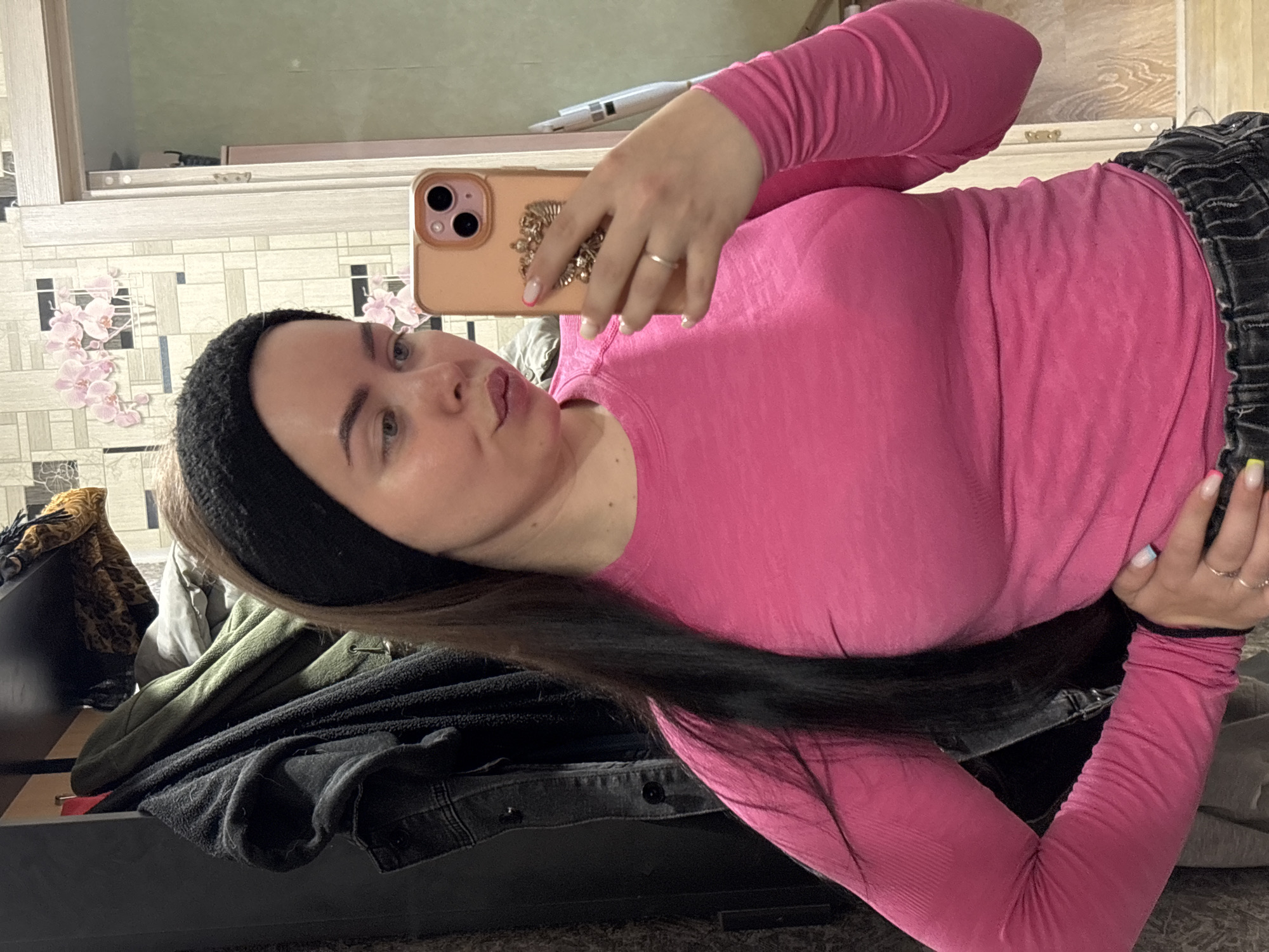 A woman is depicted taking a mirror selfie in an indoor setting, likely a home in Izhevsk, Russia. She is positioned to her right side, looking towards her reflection. She has long, dark brown hair partially covered by a black headband, and light-colored eyes. Her lips are slightly pursed in a common selfie pose.
She is wearing a bright, ribbed pink long-sleeved top and dark-colored jeans or trousers. On her left hand, she holds a light brown or gold smartphone with two visible camera lenses, framed by a case that features an intricate, metallic decorative element on the back. Her fingernails are manicured with white tips and accents of neon pink, blue, and yellow. A simple gold-colored ring is visible on her left ring finger.
The background shows a wall covered with a distinctive wallpaper featuring a geometric pattern interspersed with pink floral designs, resembling cherry blossoms. Above the wallpaper, there appears to be a light-colored wooden or laminate surface, possibly part of a cabinet or headboard. Below, a pile of clothes is visible, including a green jacket, dark fabric items, and a piece of leopard print clothing. The immediate foreground shows a dark, possibly carpeted surface. The overall impression is a casual, lived-in domestic space. No discernible text is visible in the image.