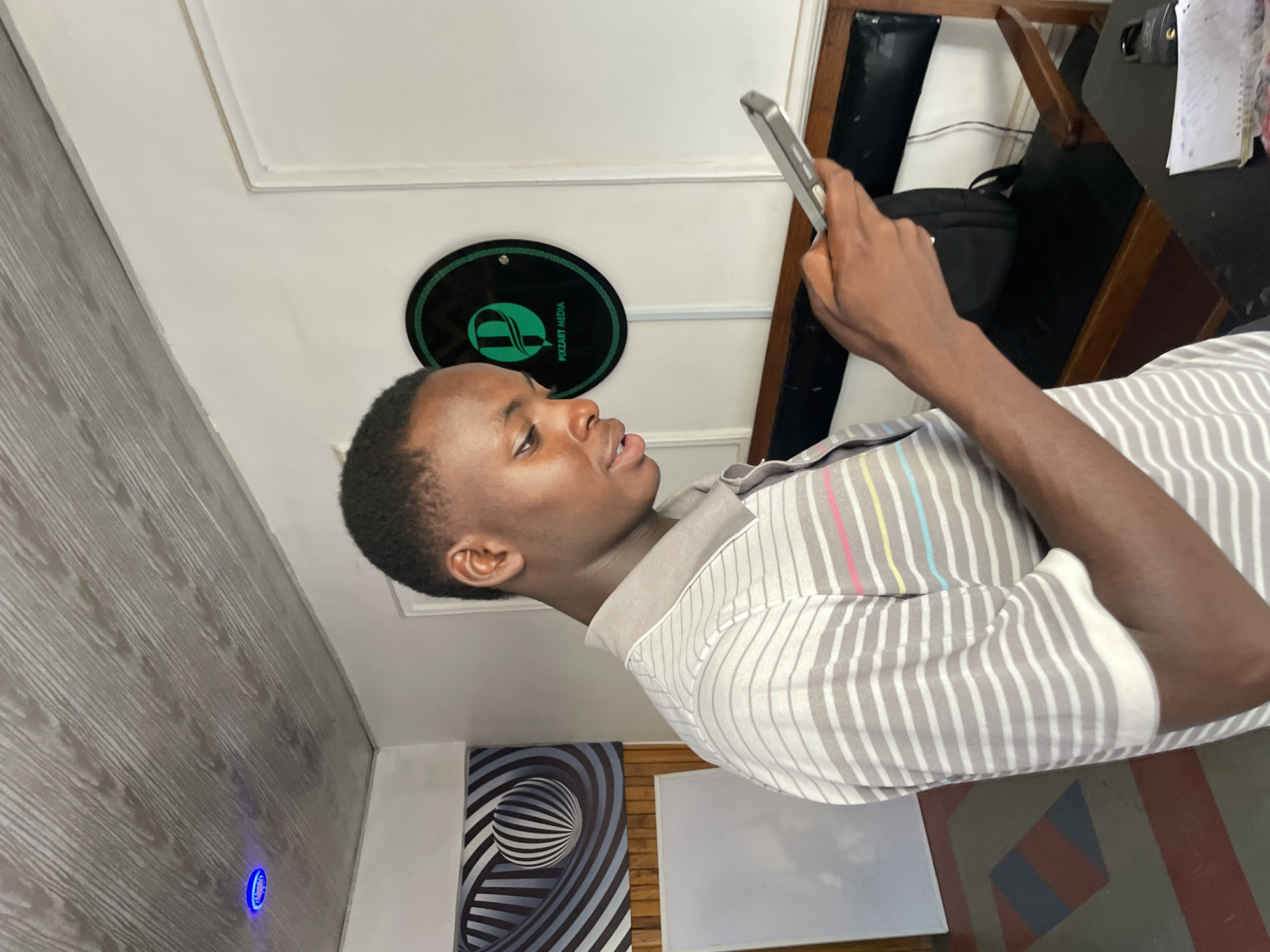 The image shows a young Black man in an indoor setting, possibly an office or a creative space. He is holding a smartphone and appears to be looking at it. He is wearing a collared shirt with a pattern of thin grey and white stripes, accented with thin colored stripes of pink, yellow, and blue on the collar area.
In the background, there is a round logo with a green design and the text "PIXZART MEDIA" written around the border. To the left of the man, the wall has a wood-like texture. Further to the left, a small blue light is visible on the wall. On the right side of the image, a desk is visible with papers and a black bag. There is also a wooden chair.
A piece of art with a spiral optical illusion featuring black and white stripes and a sphere is hanging on the wall below the "PIXZART MEDIA" logo. Another whiteboard is visible on the wall behind the man. The lighting suggests it could be daytime, though the indoor setting makes it difficult to determine the exact time of day or weather conditions. The man's expression is neutral as he focuses on his phone. The location context provided is Jos, Nigeria.