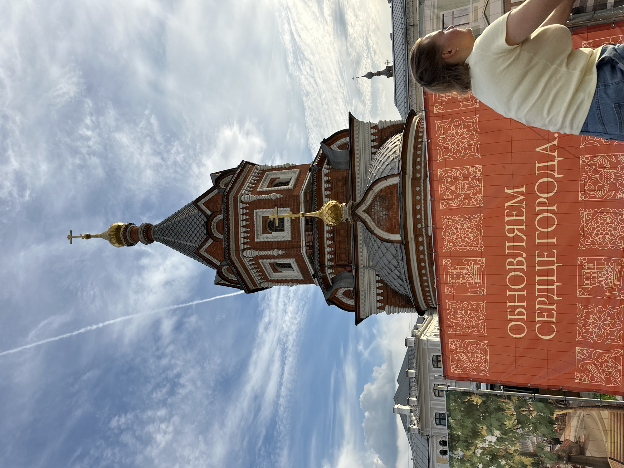 The image shows a red brick church with gold domes against a blue sky with white clouds and a contrail. In the foreground, an orange banner with decorative patterns and Russian text, "ОБНОВЛЯЕМ СЕРДЦЕ ГОРОДА" (Updating the Heart of the City), is visible. A woman with blonde hair in a light yellow t-shirt and blue jeans is partially visible on the right side of the frame, looking away from the camera. Buildings with grey roofs are seen behind the banner and to the left of the church. The scene appears to be an outdoor public space.