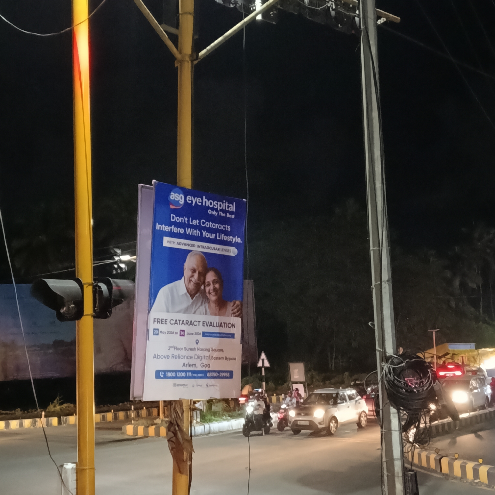 The image captures an evening scene in Arlem, Goa, near Margao, India. A street intersection is visible, with vehicular traffic consisting of cars and motorcycles. A sign promoting the "asg eye hospital" is attached to a yellow pole. The sign advertises free cataract evaluations from May 20 to June 30 at the 2nd Floor Suresh Narang Square, above Reliance Digital, Eastern Bypass, Arlem, Goa. The sign also includes two phone numbers: 1800 1200 111 and 88750-29955.