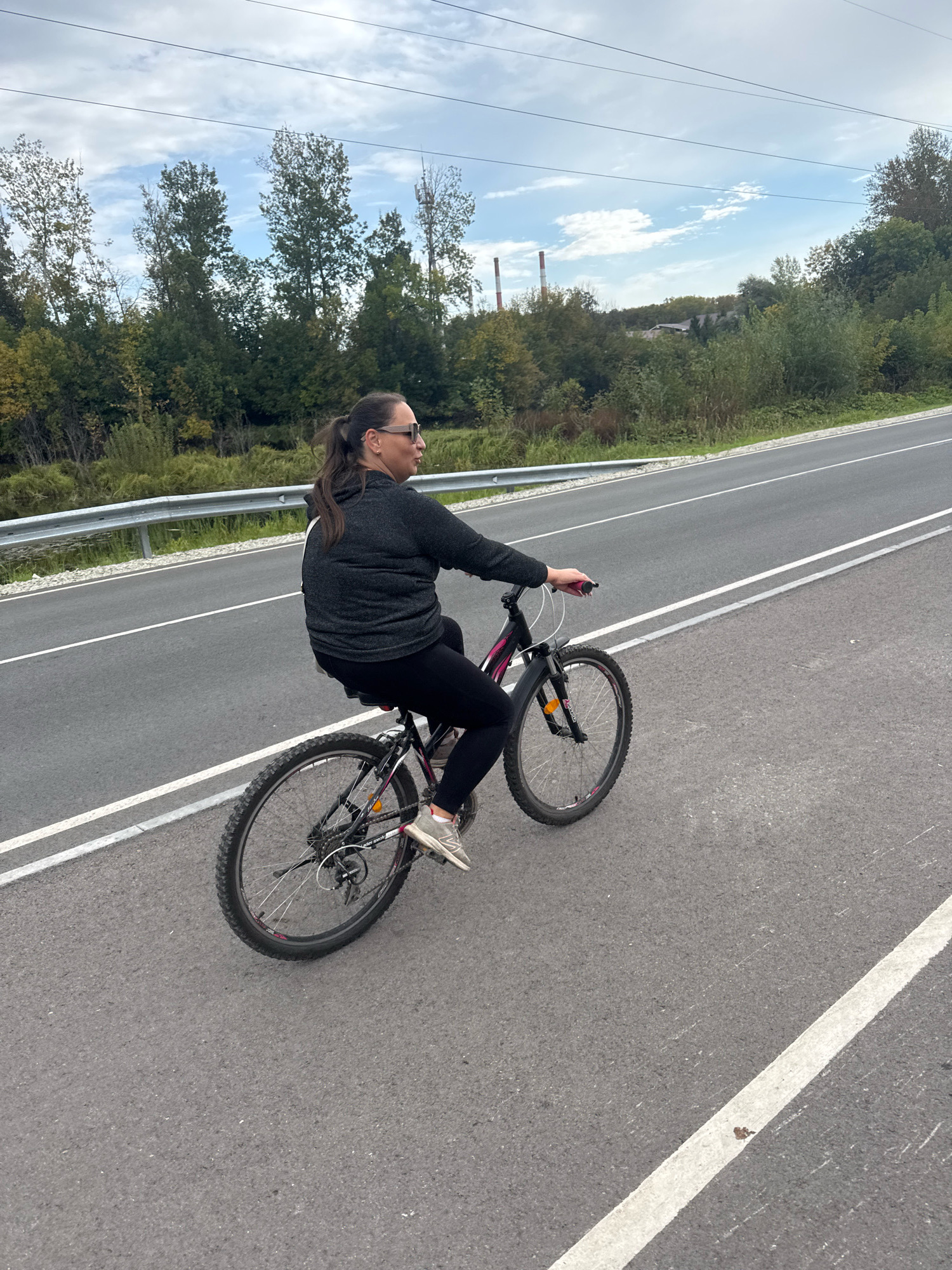 A woman is riding a bicycle on an asphalt road in Ufa, Russia. She is wearing a dark gray hoodie, black leggings, and sneakers. The bicycle is black with pink accents. The road has white lane markings. In the background, there are trees, a guardrail, and a cloudy sky with power lines and industrial chimneys.