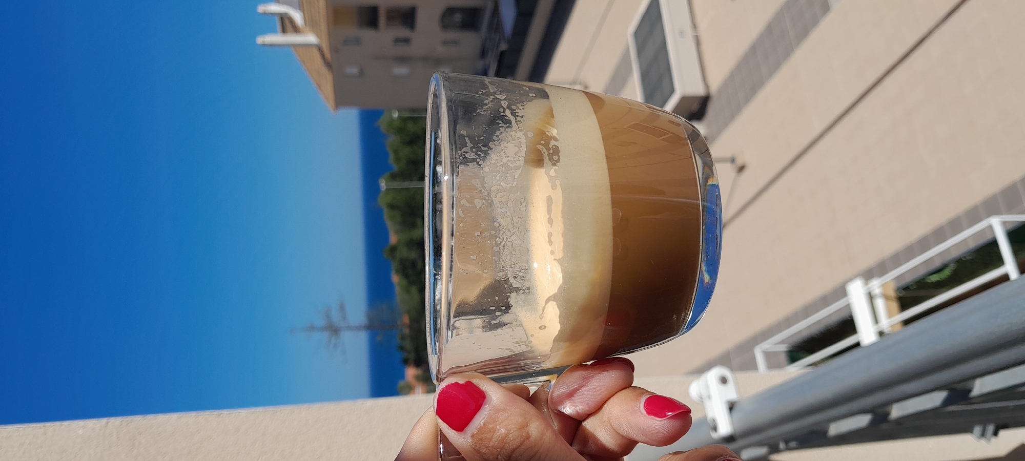 A person's hand, with red nail polish on the fingernails, holds a clear glass cup containing a layered coffee beverage, observed from a balcony in Manilva, Spain. The coffee consists of a dark brown liquid layer, a lighter brown layer above it, and a thick, creamy tan foam on top. The glass exhibits visible residue marks. Below the hand, a portion of a white and grey balcony railing is present. The background features a vibrant blue sea meeting a clear, cloudless blue sky at the horizon. Between the viewer and the sea, green foliage and trees are visible, along with a prominent, tall, leafless tree or utility pole. To the right, a multi-story building with a terracotta roof and balconies is present. The scene is brightly illuminated by natural sunlight.