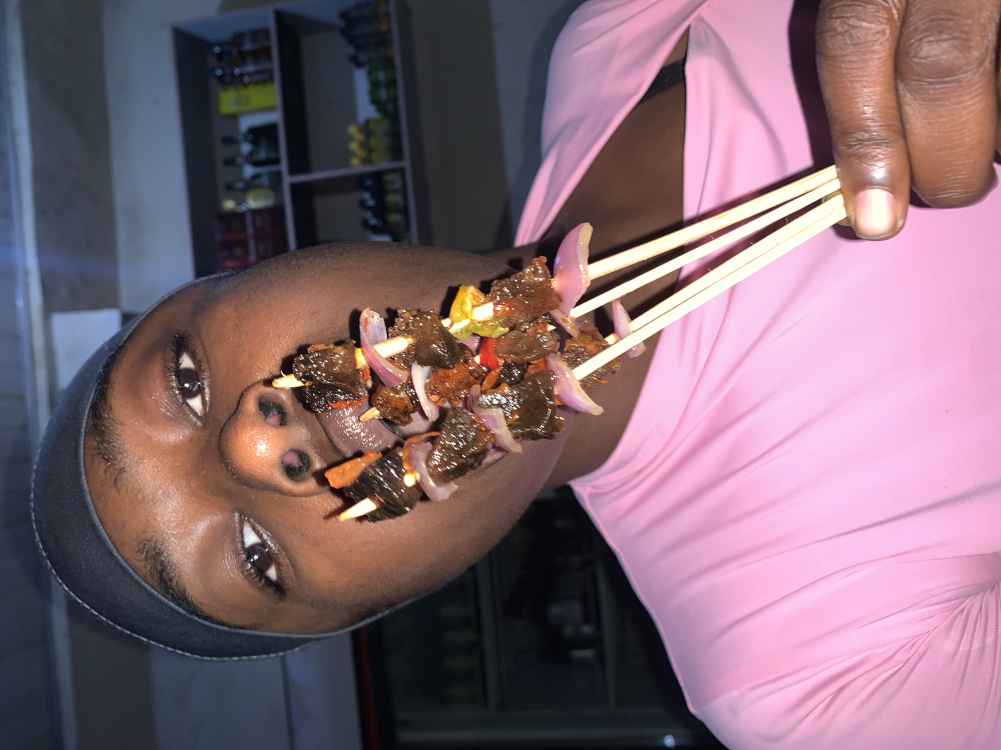 A close-up, rotated image depicts a dark-skinned person holding multiple skewers of food near their mouth. The food consists of pieces of meat with red onions. The person is wearing a pink garment and a dark head covering. The background shows a shelving unit with various objects. The location is in the city of New Stadium, Nigeria.