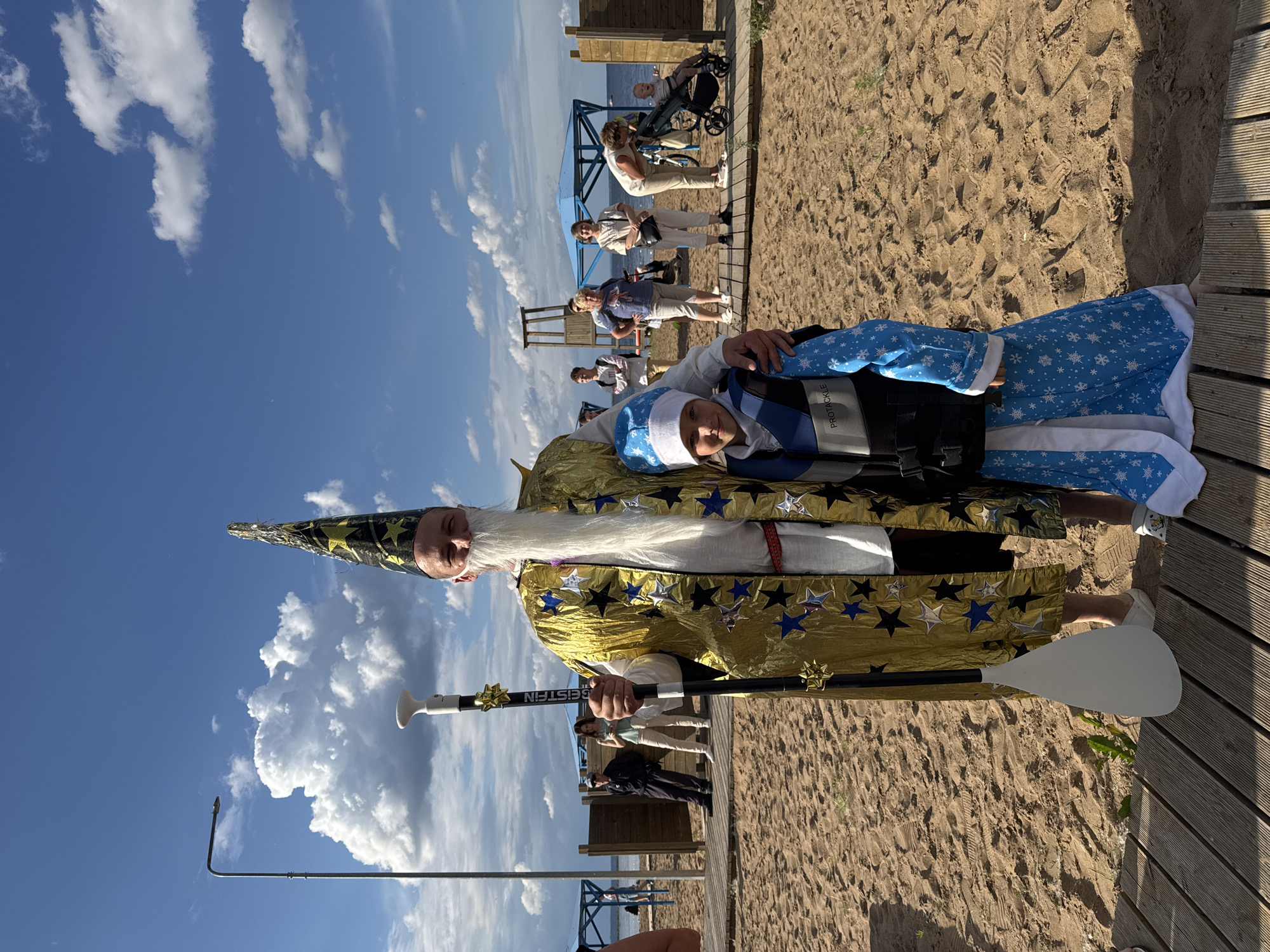 The image shows a group of people on a sandy beach near the water on a sunny day with a blue sky and scattered clouds. In the foreground, a person dressed as a wizard with a long white beard and a gold robe decorated with stars holds a paddle. Next to the wizard, another person is wearing a blue and white snowflake-patterned outfit and a blue vest.
In the background, several other people are visible, some sitting and others standing. One person is in a wheelchair. Wooden structures, possibly part of a pier or boardwalk, are present on the beach. The scene appears to be a casual gathering or event. The time of day is likely daytime, given the bright sunlight. The location is identified as Chaikovsky, Russia. The word "GEISTEIN" is visible on the paddle.