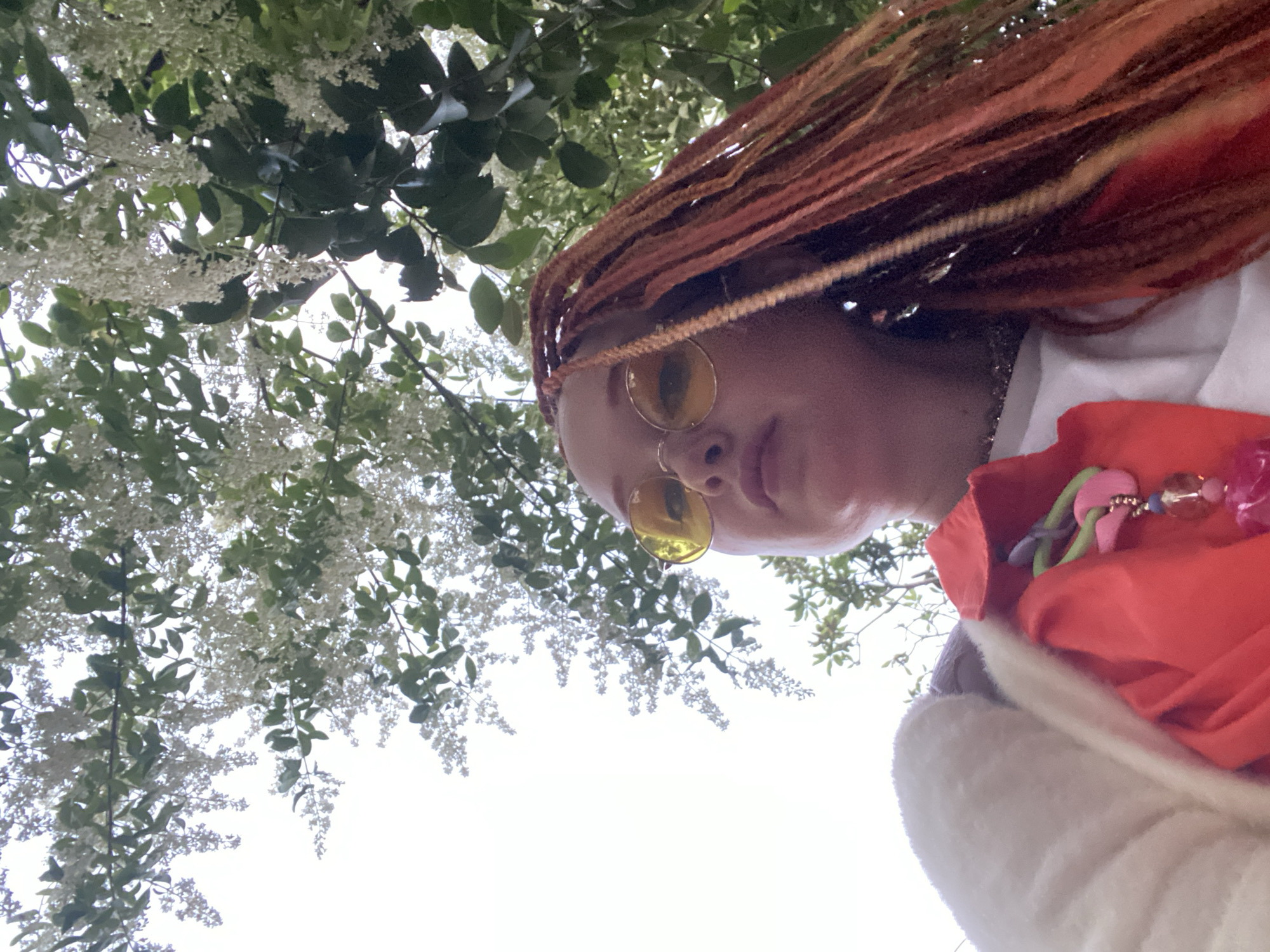 The image is a low-angle shot looking upwards, capturing a person against a backdrop of flowering trees and a bright sky. The subject is a person with reddish-orange dreadlocks or braids, wearing round, yellow-tinted sunglasses. Their face is partially visible, looking down towards the camera with a neutral expression. They appear to be wearing a white top or shirt, and on the right side of the frame, there's a vibrant orange fabric, possibly a scarf or part of an accessory, adorned with colorful beads and charms in shades of pink, green, and blue. A soft, light-colored material is visible beneath the orange fabric.
The upper and left sections of the image are filled with branches of a tree or shrub, densely covered with small white blossoms and green leaves. The bright, clear or lightly overcast sky is visible through the foliage, indicating daytime. The perspective suggests the photograph was taken from below the subject, looking up at them as they stand beneath the blooming canopy. The setting is outdoors, likely in a park or garden in Sochi, Russia, during a period of spring or early summer when such trees are in full bloom. There is no visible text in the image.