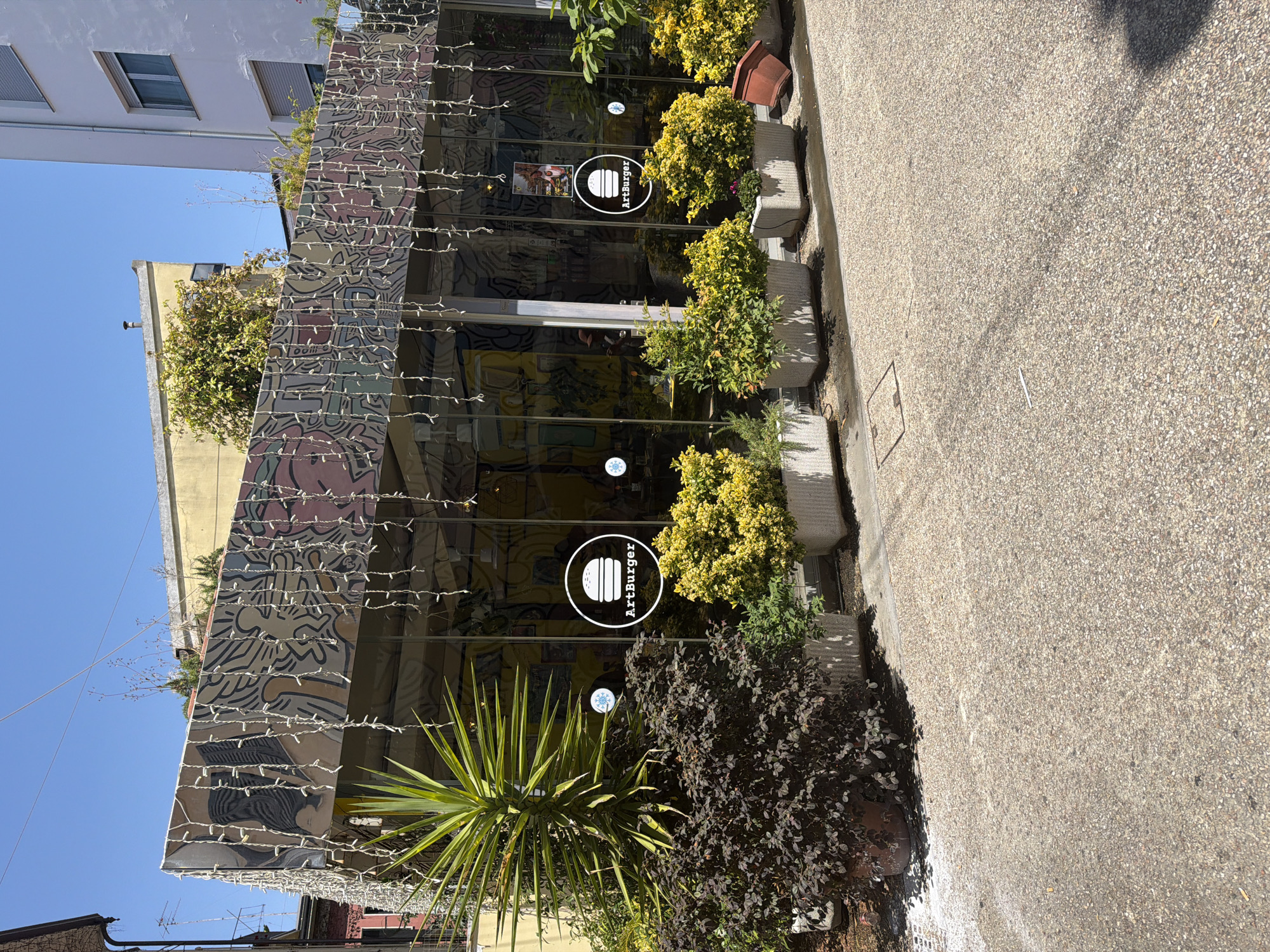 An exterior, ground-level, slightly angled medium shot captures a modern building with a glass facade and an artistic, mural-covered upper story adorned with hanging string lights. The building appears to be a restaurant or cafe, as indicated by a circular logo on the glass featuring a hamburger icon and the text "ArtBurger." In front of the building, a row of potted plants, including a large yucca-like plant and bushy yellow-flowering plants, are arranged along the glass, creating a lush, green barrier. The ground in front of the building is paved with a speckled, aggregate material. In the background, parts of adjacent buildings are visible, one painted yellow and another white, suggesting an urban setting. The sky above is clear and blue. The scene is bathed in bright sunlight, casting shadows on the ground and highlighting the textures of the pavement and vegetation. There is no discernible activity of people in the image.
