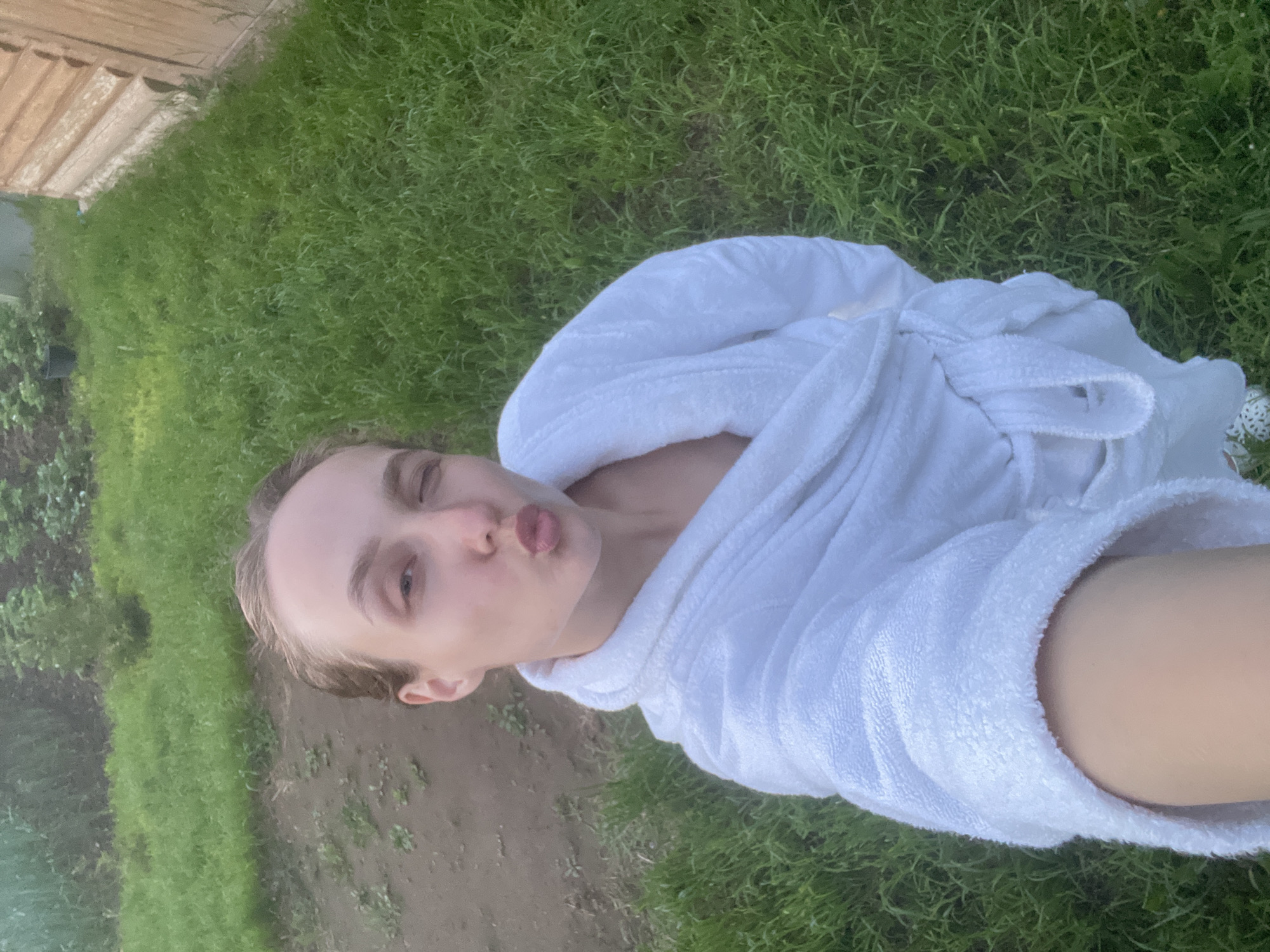 A young individual is captured in a sideways selfie outdoors, facing left. The person has light brown hair and fair skin, and is wearing a white, fluffy terry cloth bathrobe, which is tied at the waist. They are making a playful expression with pursed lips as if blowing a kiss, and one eye is squinted or winking.
The background of the image features a green, grassy area with patches of bare soil. In the upper left of the rotated image, there is denser green foliage, resembling a hedge or bushes, and a light-colored wooden fence or structure. The lighting suggests it is daytime, possibly late afternoon or early evening, with soft, natural light indicating fair weather. The setting appears to be a backyard or garden in Sharkan, Russia. The overall mood conveyed by the subject's expression is playful and relaxed. There is no visible text in the image.