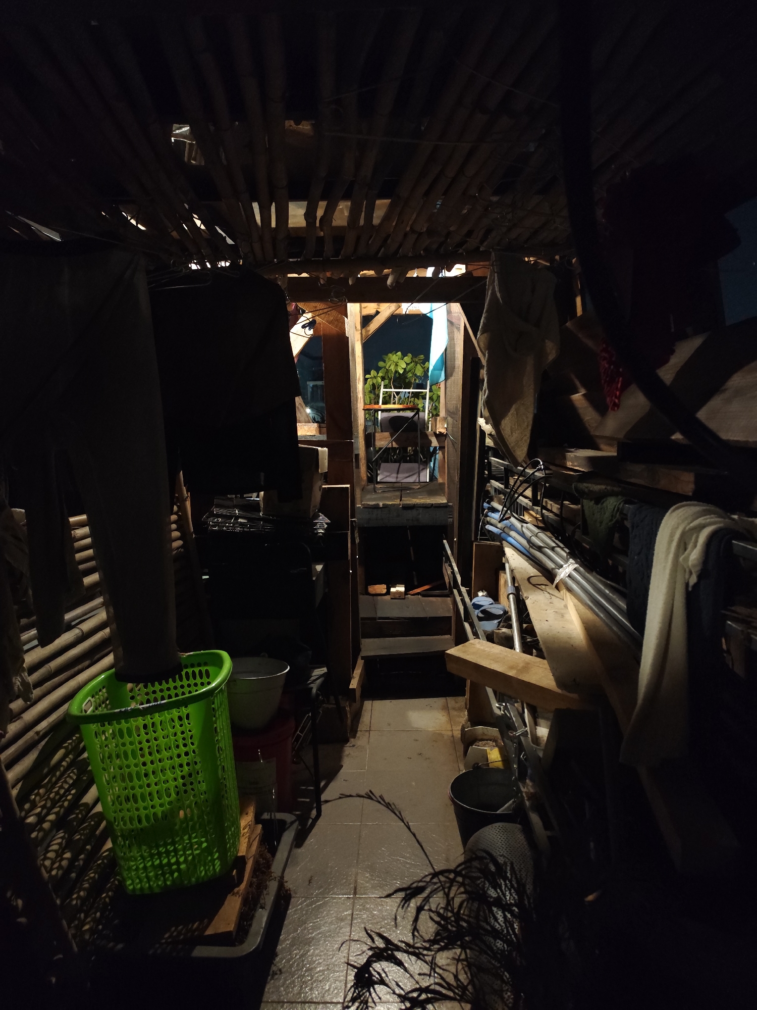 The image depicts a cluttered interior space in Da Lat, Vietnam. The roof is constructed of bamboo. The space is filled with assorted objects including lumber, pipes, buckets, and a green plastic laundry basket. A small staircase leads to an outdoor area with a table, chairs, and potted plants. The overall lighting is dim, with a brighter area visible in the outdoor space. The scene appears to be a storage area or workshop.