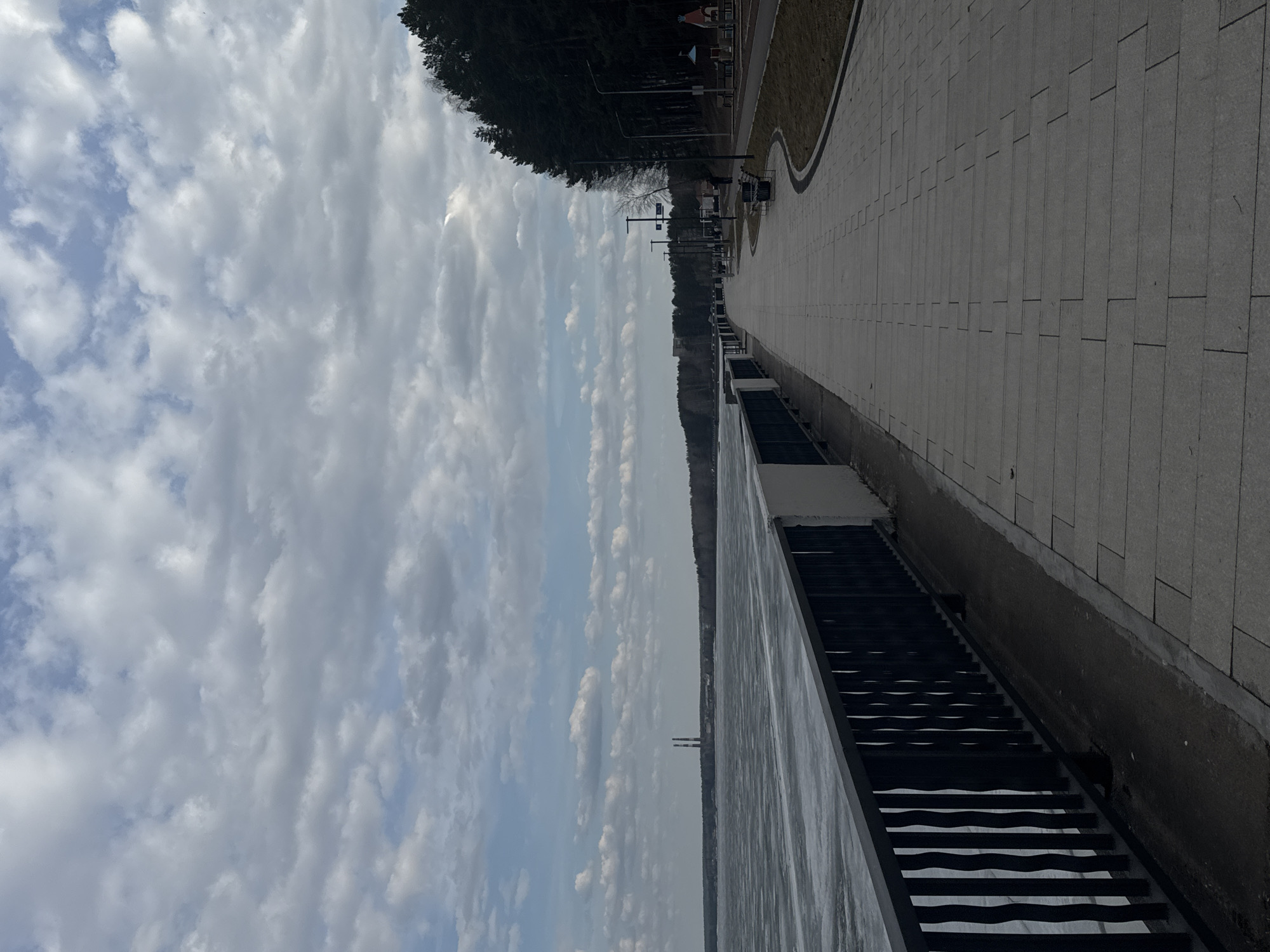 The image displays a waterfront embankment and pathway under a partly cloudy sky, likely in Chaikovsky, Russia. The perspective is from an elevated position looking parallel to the water.
In the upper portion of the frame, the sky is filled with numerous white and grey cumulus clouds, with patches of clear blue sky visible between them.
On the right side, a wide, paved promenade or walkway, constructed from light-colored rectangular tiles, stretches into the distance. To the right of the walkway, a narrow strip of green grass curves alongside it, backed by a dense line of dark evergreen trees. Several modern streetlights and benches are spaced along the pathway.
On the left side, a large body of water, possibly a river or reservoir, extends across the mid-ground. The water's surface appears calm and partially or fully covered with ice, reflecting the sky. A dark, wide set of stairs descends from the embankment structure into the water.
In the far distance, across the body of water, a low horizon of land is visible. Above this distant land, two tall, slender, light-colored industrial chimneys or towers rise prominently against the sky.
The scene depicts a calm daytime environment, possibly in late winter or early spring, given the icy water. No people or specific activities are visible. There is no visible text in the image.