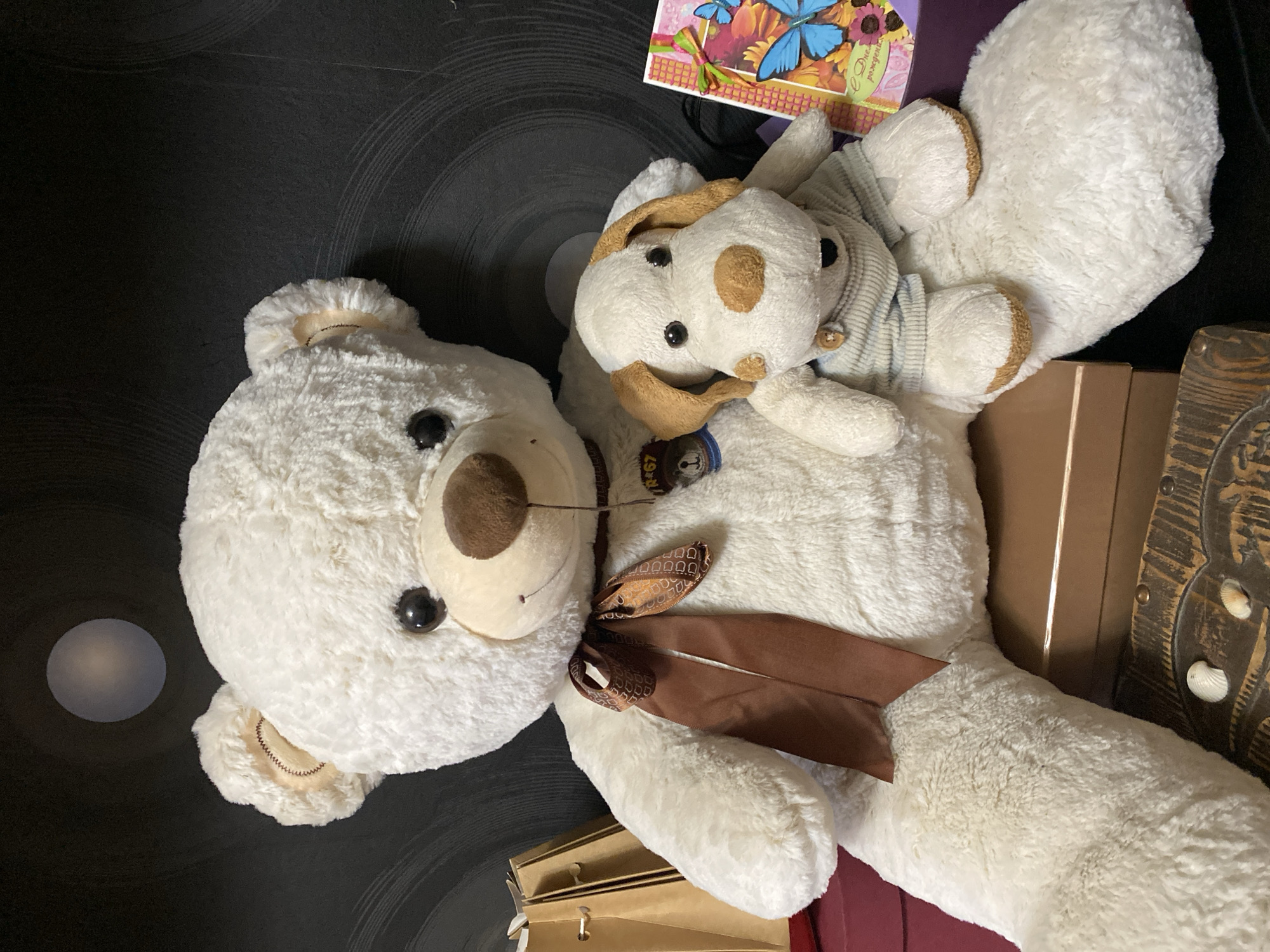 The image depicts two stuffed animals, a large white teddy bear and a smaller stuffed dog, positioned in the foreground. The teddy bear is seated and faces forward, with a brown ribbon tied around its neck. The stuffed dog is sitting in front of the teddy bear's left arm. Behind the stuffed animals, several items are visible, including two brown paper bags, a gift box, and a greeting card with blue butterflies and flowers. The background features a dark wall with circular patterns and a faint, circular light source. The overall scene suggests an indoor setting, possibly a home. This image was taken in Novaya Usman', Russia.