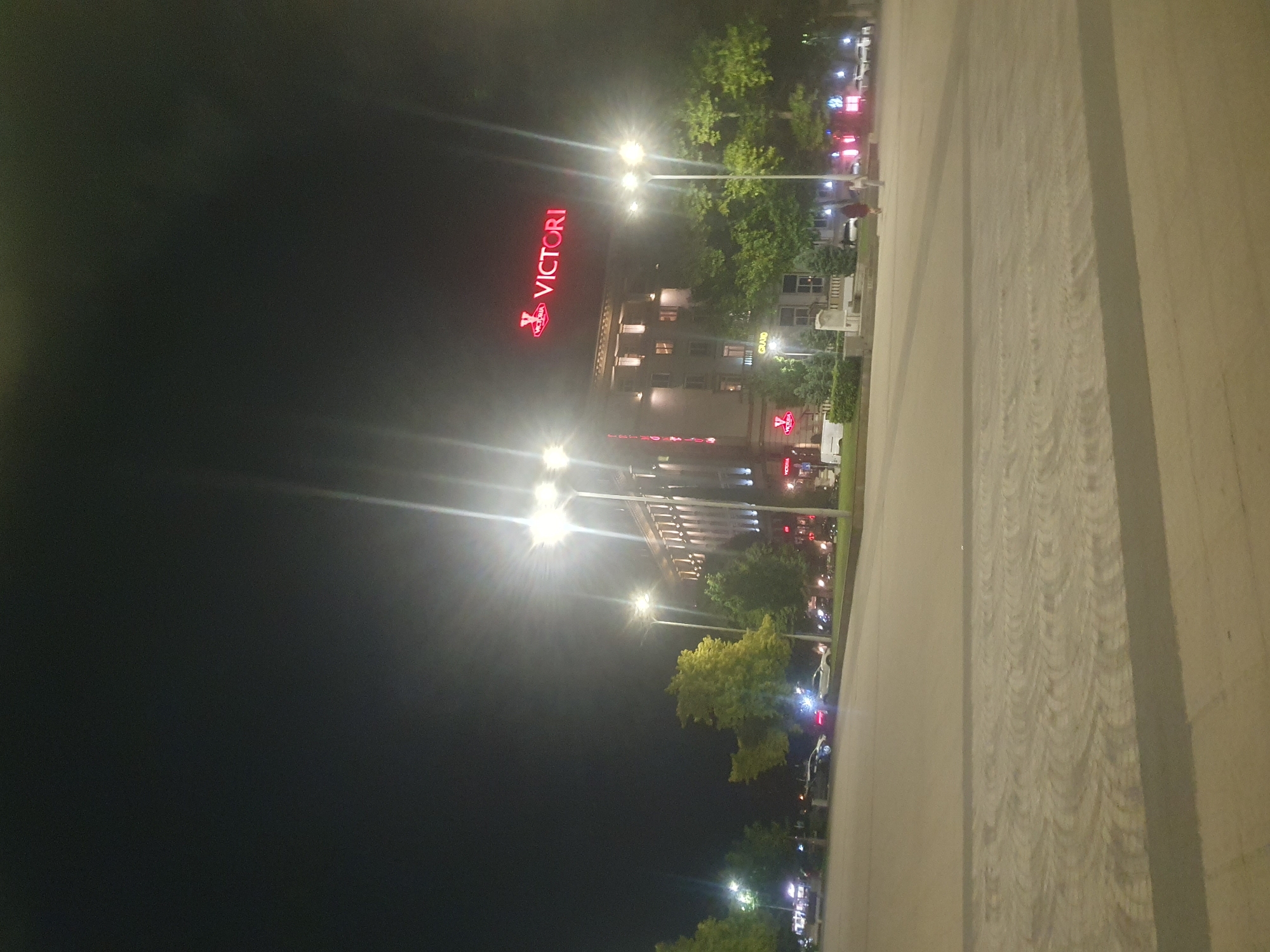 The image depicts a night scene in Plovdiv, Bulgaria, featuring the Victoria Grand Hotel. The hotel is illuminated with lights, and its name "VICTORIA" is displayed in red lettering on the facade. Streetlights line the road in front of the hotel, with trees and other urban greenery visible between the streetlights and the building. A paved area, potentially a sidewalk or plaza, takes up the foreground of the image. The sky is dark, suggesting a late hour.