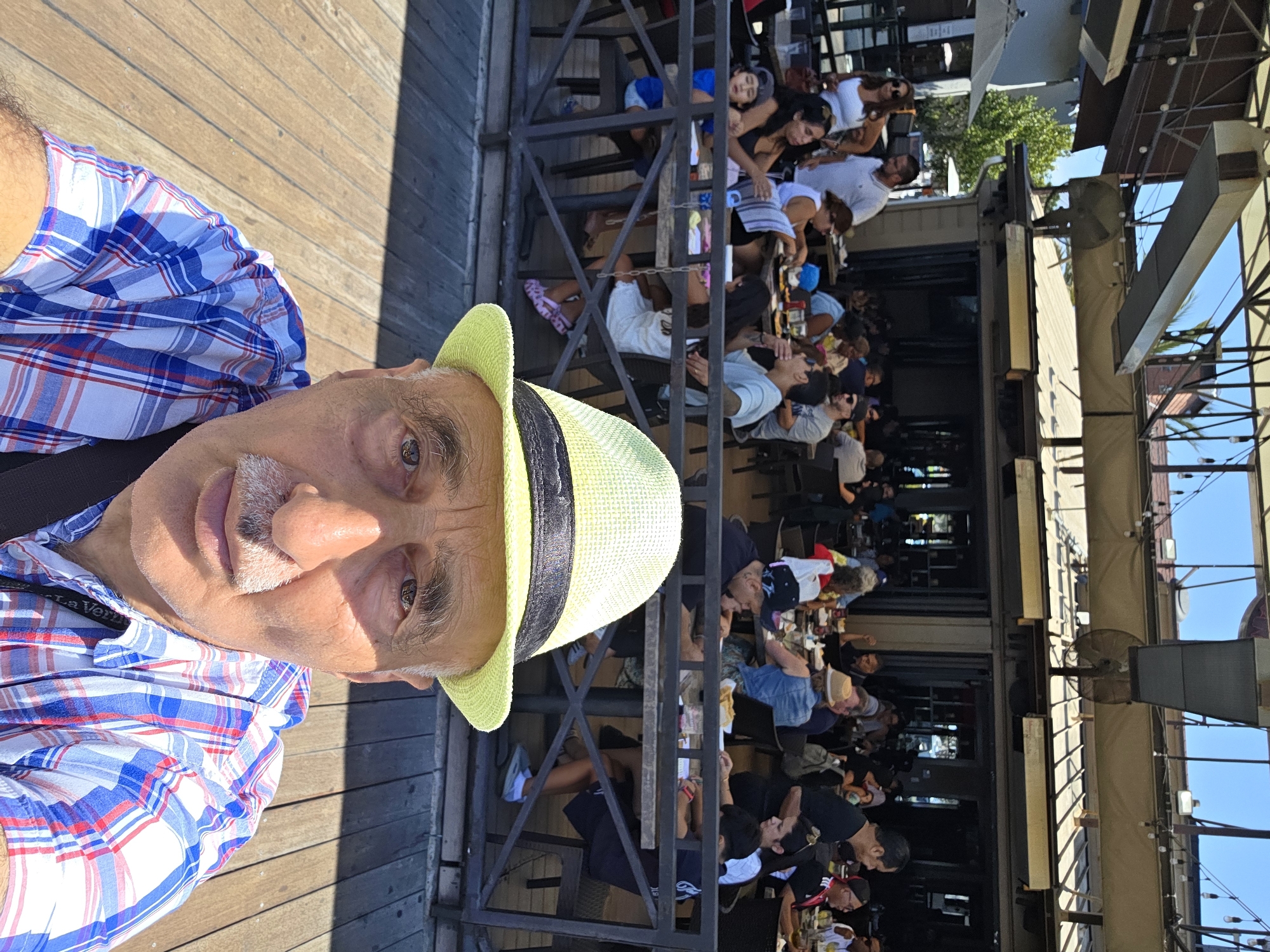 The image shows a man in the foreground wearing a light green hat and a blue and white plaid shirt. He is taking a selfie outdoors, with a busy outdoor restaurant patio in the background. The restaurant appears to be located on a wooden deck, possibly by the water, suggesting a location in Long Beach, USA, known for its waterfront dining. The patio is filled with people seated at tables, some eating and socializing, while others are interacting with staff or looking around. The architecture of the restaurant is visible, with a covered roof and open-air seating areas. The overall atmosphere suggests a sunny day with a lively, casual dining experience.