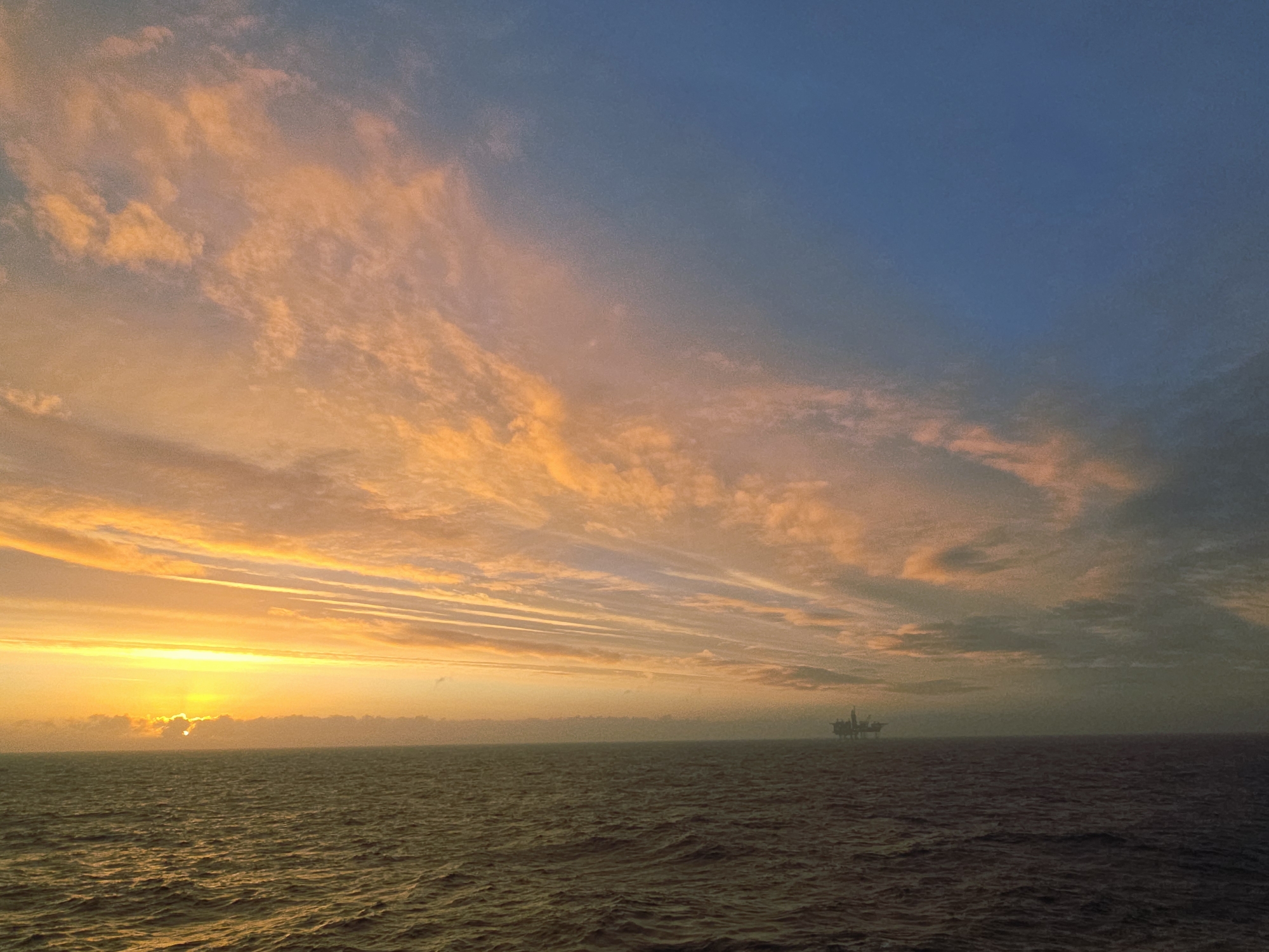The image depicts an ocean scene at sunset. The sun is low on the horizon, obscured by clouds, casting a golden glow across the water and the lower part of the sky. The sky above transitions from orange hues near the horizon to a bluish color in the upper-right corner. Clouds are scattered throughout the sky, varying in density and color. In the distance, a silhouette of what appears to be an oil rig is visible on the horizon. The ocean itself is dark and undulating, reflecting the colors of the sky. The location is the ocean.
