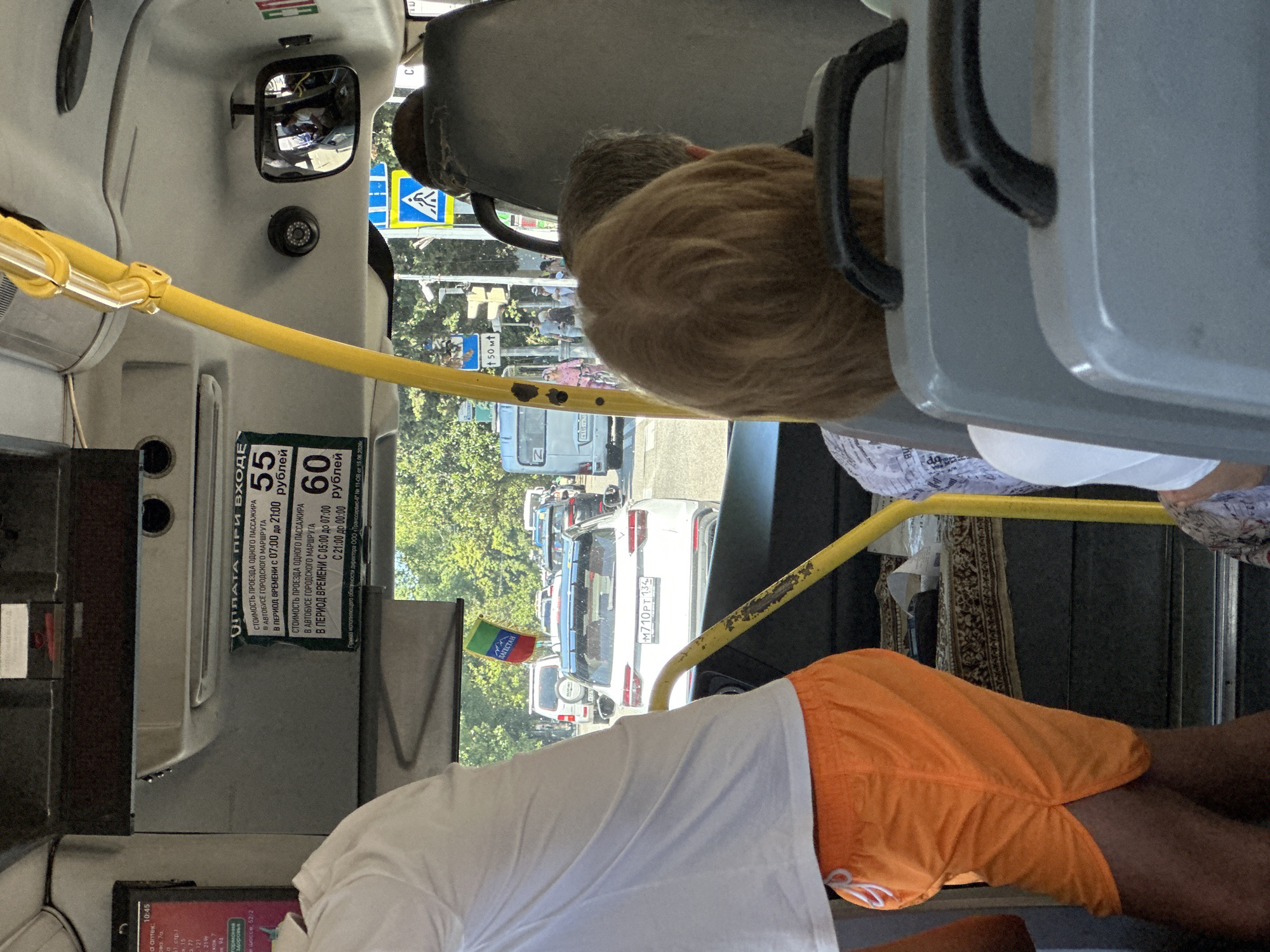 The image shows the interior of a bus, likely in Sochi, Russia, as evidenced by the signage with Cyrillic script and the context of the region. The scene captures a moment of transit with several passengers visible. In the foreground, a person wearing an orange-colored athletic short is standing, partially obscuring the view of the seats and other passengers. Behind them, other passengers are seated.
The bus interior features a yellow pole, a common fixture in public transportation for passengers to hold onto. The ceiling of the bus is visible, with some overhead compartments. A mirror mounted on the ceiling reflects parts of the interior, including a security camera.
Outside the bus, through the windows, a street scene unfolds. Cars are in traffic, indicating a busy urban environment. A pedestrian crossing sign is visible on the right side of the street, suggesting a well-trafficked area. Trees and buildings can be seen in the background, typical of a city landscape.
A prominent sign inside the bus displays information about fare prices, written in Russian. The sign indicates the cost of a single passenger ride on a city bus, with different prices for different times of the day. Specifically, it shows a fare of 55 rubles for rides between 07:00 and 21:00, and 60 rubles for rides between 05:00 and 07:00, and again from 21:00 to 00:00. This detail further supports the location as Russia.
A small flag, with green, blue, and red colors, is hanging from the interior of the bus near the front. The flag's design suggests it may represent a region or ethnic group within Russia. The overall impression is of an ordinary day on a public bus, navigating through city traffic.