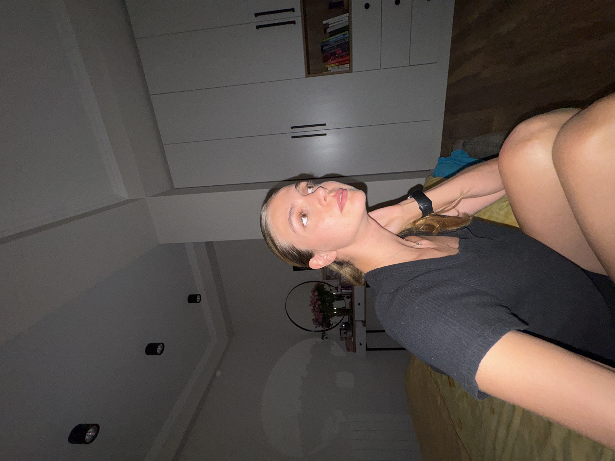 The media file displays a young woman posing indoors, captured in what appears to be a selfie or a close-up photo. The image is initially rotated 90 degrees counter-clockwise.
When viewed correctly (rotated 90 degrees clockwise), the woman is positioned in the lower right and central foreground, lying down on her side or back, looking upwards towards the upper left. She has light-colored hair pulled back, light eyes, and is wearing a black ribbed short-sleeved top and a black wristwatch on her left wrist. A delicate silver necklace with a small pendant is visible around her neck. Her expression is neutral and contemplative. Her bent legs are visible in the upper right part of the frame, resting on a textured, yellow or mustard-colored surface, likely a bed or couch.
The background is an interior space, probably a bedroom or living area. On the upper left, a large, light grey or white modular cabinet system with black horizontal handles dominates the background. One central section of the cabinet is open, revealing a stack of colorful books. To the lower left, a wall features several recessed black spotlights. Further back and slightly to the left of the woman, a shelf holds a vase of pink flowers behind a large, dark circular decorative hoop. A dark wooden floor is visible in the upper right background. The overall lighting is dim, suggesting evening or nighttime, illuminated by artificial light from the ceiling spots. There is no visible text in the image.