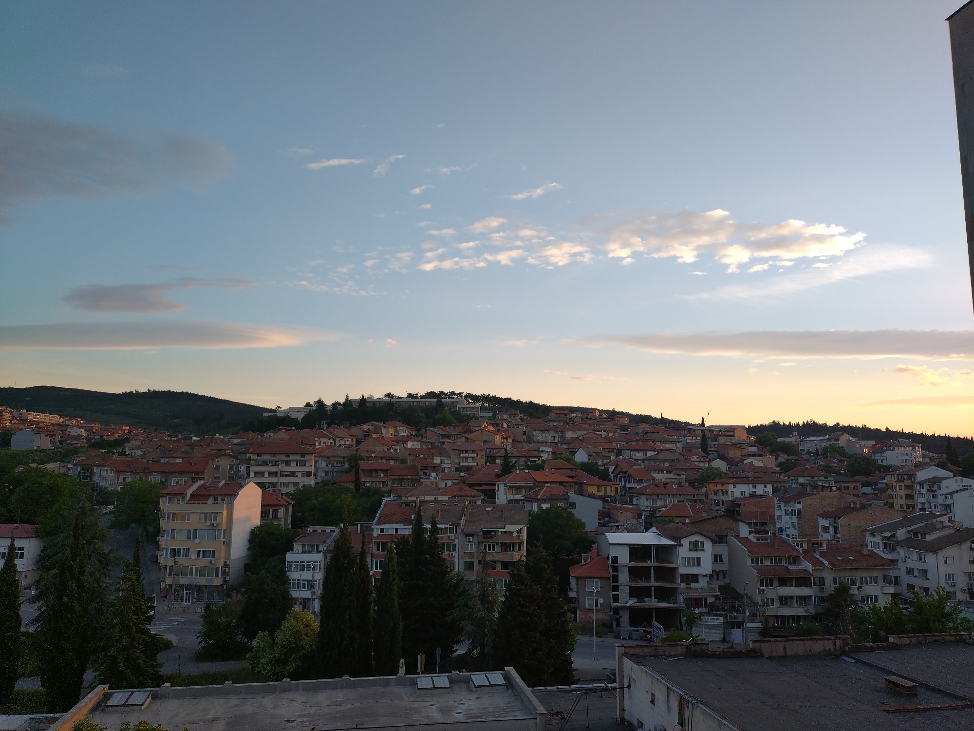A cityscape view from an elevated perspective at twilight in Stara Zagora, Bulgaria. The foreground features rooftops of buildings, with several tall, dark green trees visible. Mid-ground is occupied by densely packed residential buildings with orange-tiled roofs, interspersed with green trees and varied architectural styles. A gentle slope leads uphill, topped by more buildings silhouetted against a brightly lit sky. The sky is blue with tinges of orange and pink, especially near the horizon, and scattered with thin clouds.