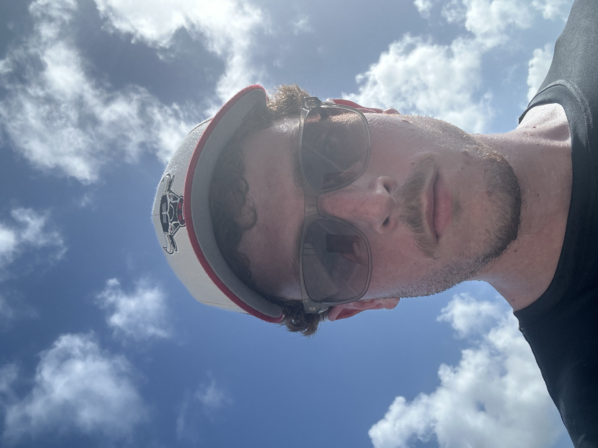 A low-angle selfie shows a person wearing a baseball cap and sunglasses. The person has a beard and is wearing a black shirt. The background is a blue sky with scattered white clouds. The location cannot be determined.