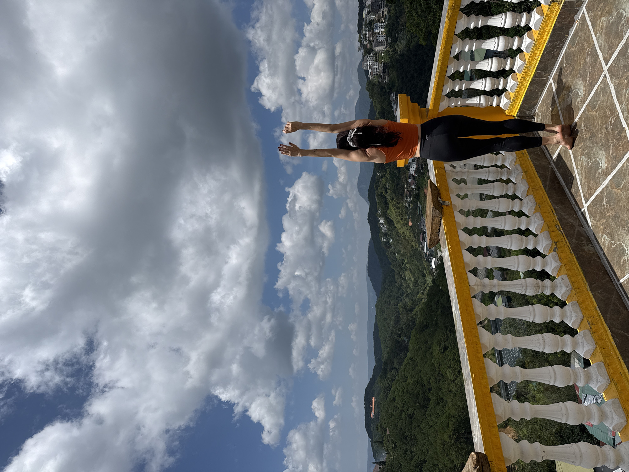 A woman stands on a balcony overlooking a verdant valley in Da Lat, Vietnam. She is facing away from the camera, stretching her arms upwards towards a cloudy blue sky. The balcony features a white balustrade with yellow accents and a tiled floor. The landscape beyond is a tapestry of rolling, tree-covered hills dotted with buildings. The clouds are a mix of bright white cumulus and darker, more ominous formations.