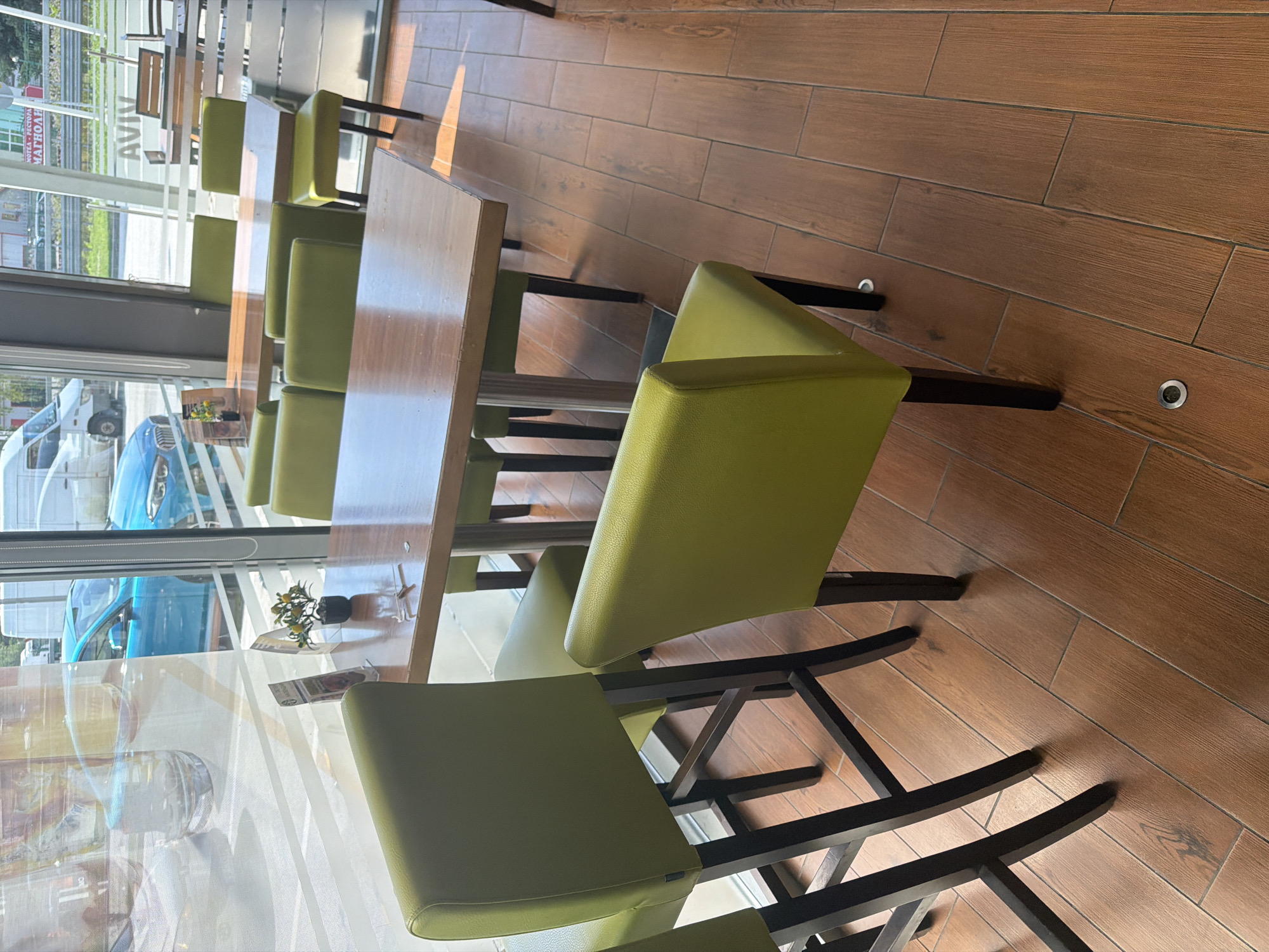 An interior shot shows a clean and empty restaurant or cafe setting. The scene features rows of tables with matching green upholstered chairs, which have dark, possibly wooden or metal, frames. The tables are rectangular with light brown wooden tops. The floor is covered with wood-look ceramic tiles in varying shades of brown, and small circular embedded lights are visible in the floor.
A large window dominates the left side of the image, offering a view of the outside. Through the window, several vehicles are visible, including a bright blue car and multiple white vans, suggesting a street or parking area. Reflections on the window obscure some of the outdoor view and also show parts of the interior. On one of the tables near the window, a small decorative plant in a black pot is placed.
Sunlight streams into the room, creating bright spots and shadows, indicating it is daytime. On an outdoor sign visible through the window, partially obscured text includes "AVIV" and parts of other words like "HOTEL PECTOPA" (likely meant to be "HOTEL РЕСТОРАНТ" or "HOTEL RESTAURANT") and "МАГНОАЛ". No people are present in the scene. The overall impression is a well-lit, organized dining area awaiting customers in Borovo, Bulgaria.