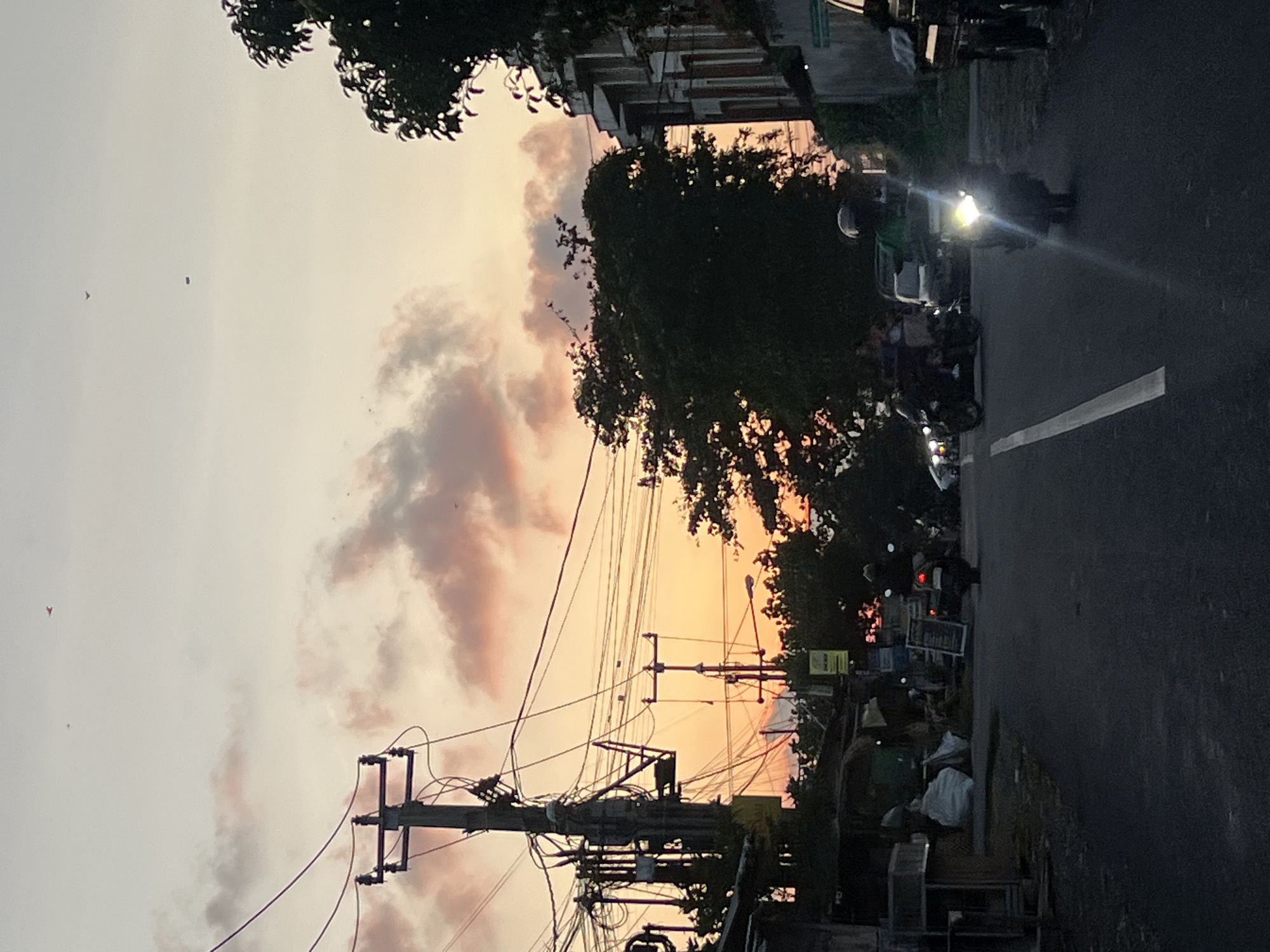 This is a vertical, medium-distance, eye-level shot of a street in Denpasar, Indonesia, during twilight. The sky is a gradient of orange and pink, with scattered clouds. Several telephone poles with a dense network of wires dominate the left side of the frame, casting shadows on the street below. A few buildings, some with white walls and red-tiled roofs, are visible on the right side. A dark-colored car is parked on the right edge of the image. Several motorcycles are present on the road, some in motion and others parked. One motorcycle in the foreground has its headlights on, casting a bright glare. The road is asphalt with a white line marking its center. Vegetation, including trees, can be seen throughout the scene. A few birds are flying in the sky. There are some small structures and debris visible on the left side of the street.