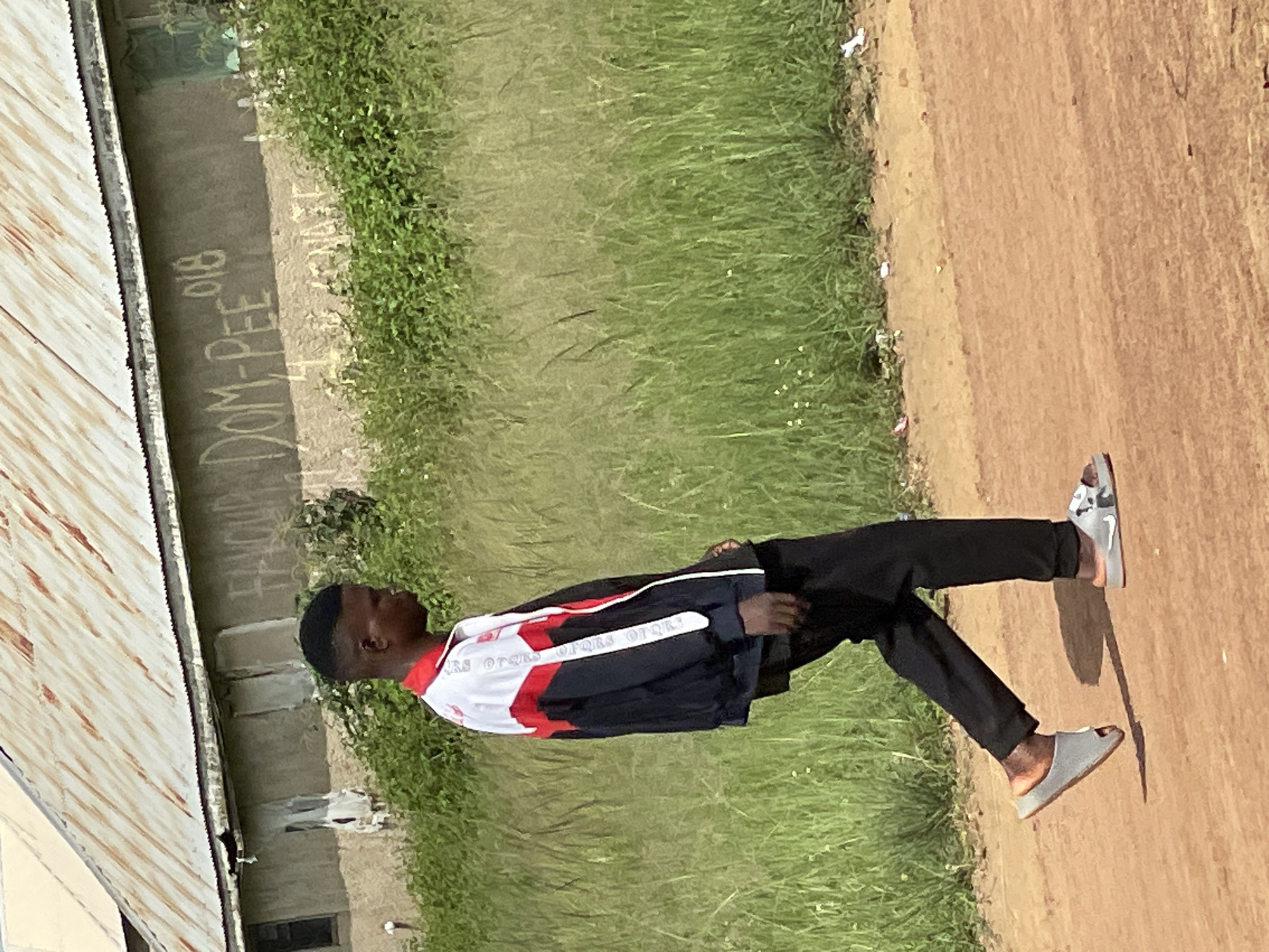 A young Black male, with a fade haircut, walks on a dirt road in Durumi, Nigeria. He is wearing a black, red, and white track jacket, black pants, and two different styles of slides on his feet. The left foot has a grey slide with a cutout, and the right foot has a white slide with black accents. Behind him, a weathered building with a corrugated metal roof stands adjacent to a patch of tall, green grass and vegetation. Graffiti, including the words "FAVOUR DOM-PEE 018," is visible on the building's wall. Debris is scattered on the roadside. The overall scene depicts an outdoor, somewhat neglected urban environment.