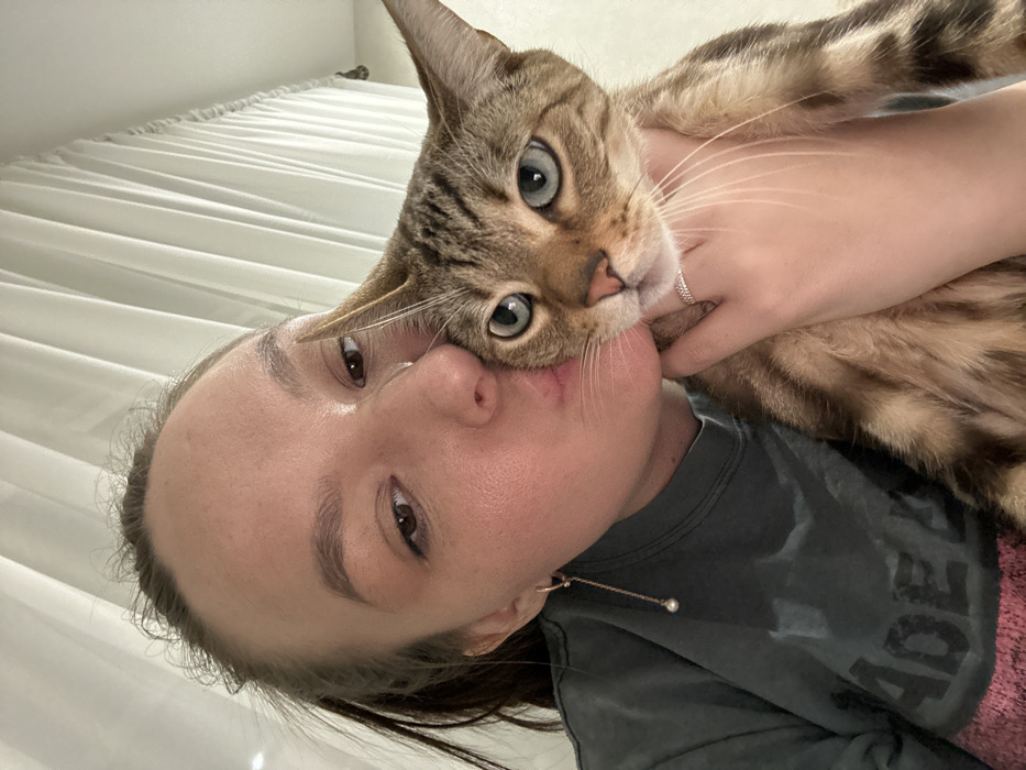 An adult female, with light skin and dark hair pulled back, is depicted in a close-up indoor photograph, embracing a domestic cat. Her left cheek is pressed against the cat's head, and her lips are pursed towards it. She wears a dark grey t-shirt, a gold-colored dangling earring with a pearl on her left ear, and a thin ring on the ring finger of her right hand, which is supporting the cat. The cat, a tabby breed exhibiting brown, tan, and black striped fur and light blue-green eyes, is positioned facing forward. White, vertically pleated curtains form the background. This scene is situated in the city of Chaikovsky, Russia.