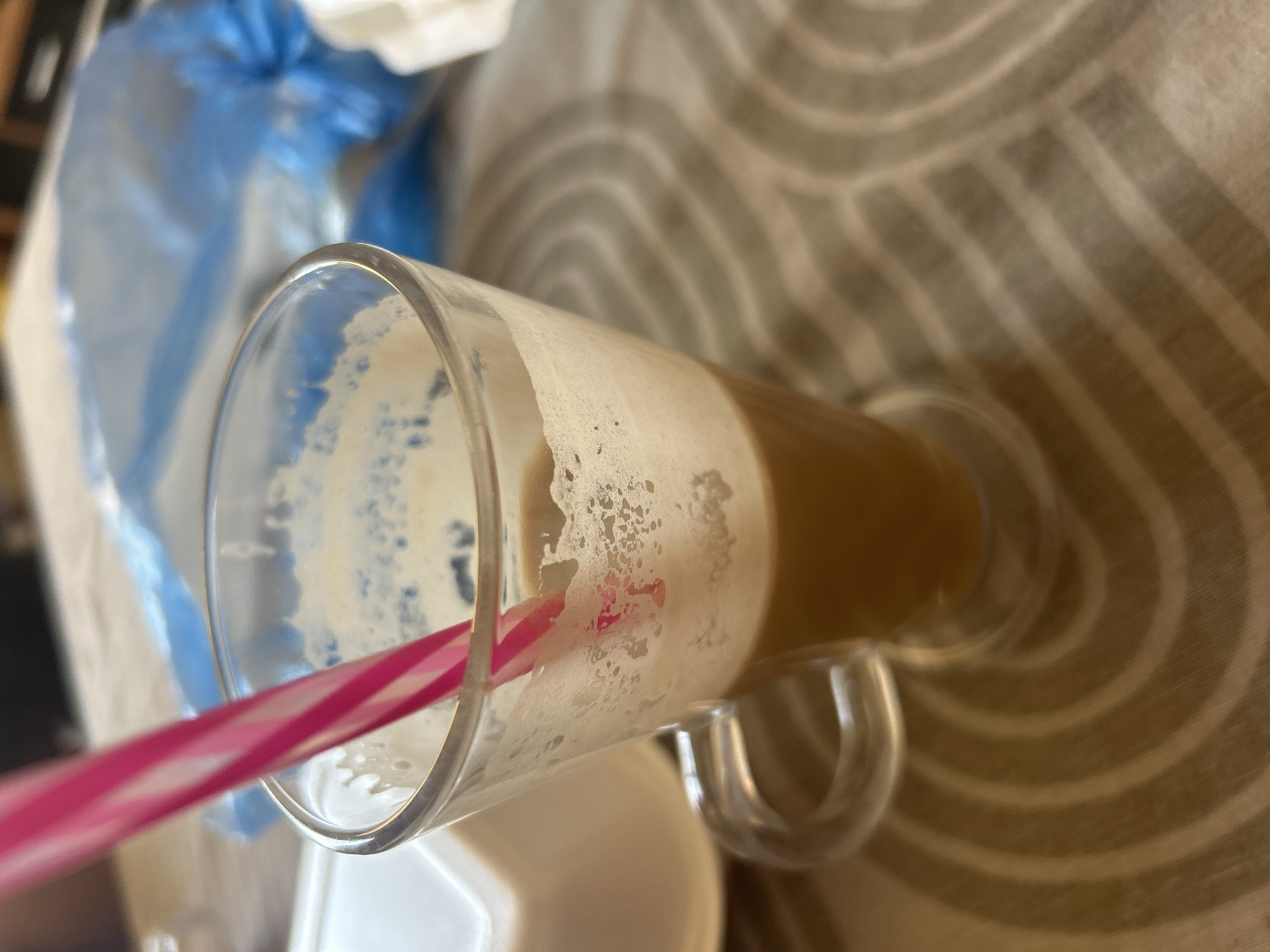 The image shows a close-up of a glass mug filled with a frothy, light brown beverage, likely coffee. A pink and white striped straw extends from the mug. The mug has a clear glass handle and rests on a table with a textured beige and white striped tablecloth. In the background, out of focus, is a white object on the left and a crumpled blue plastic bag on the right. The scene is indoors, suggesting a cafe or restaurant setting. There is no discernible information about the location being in Plovdiv, Bulgaria, or any specific city.